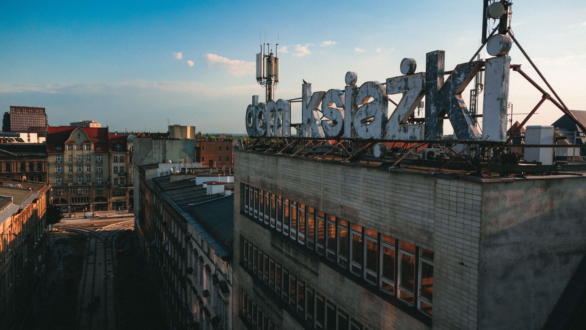 Widok z góry na budynek z dużym, metalowym napisem 'dom Książki' na dachu, z antenami i widokiem na miasto w tle, w ciepłym świetle zachodzącego słońca.