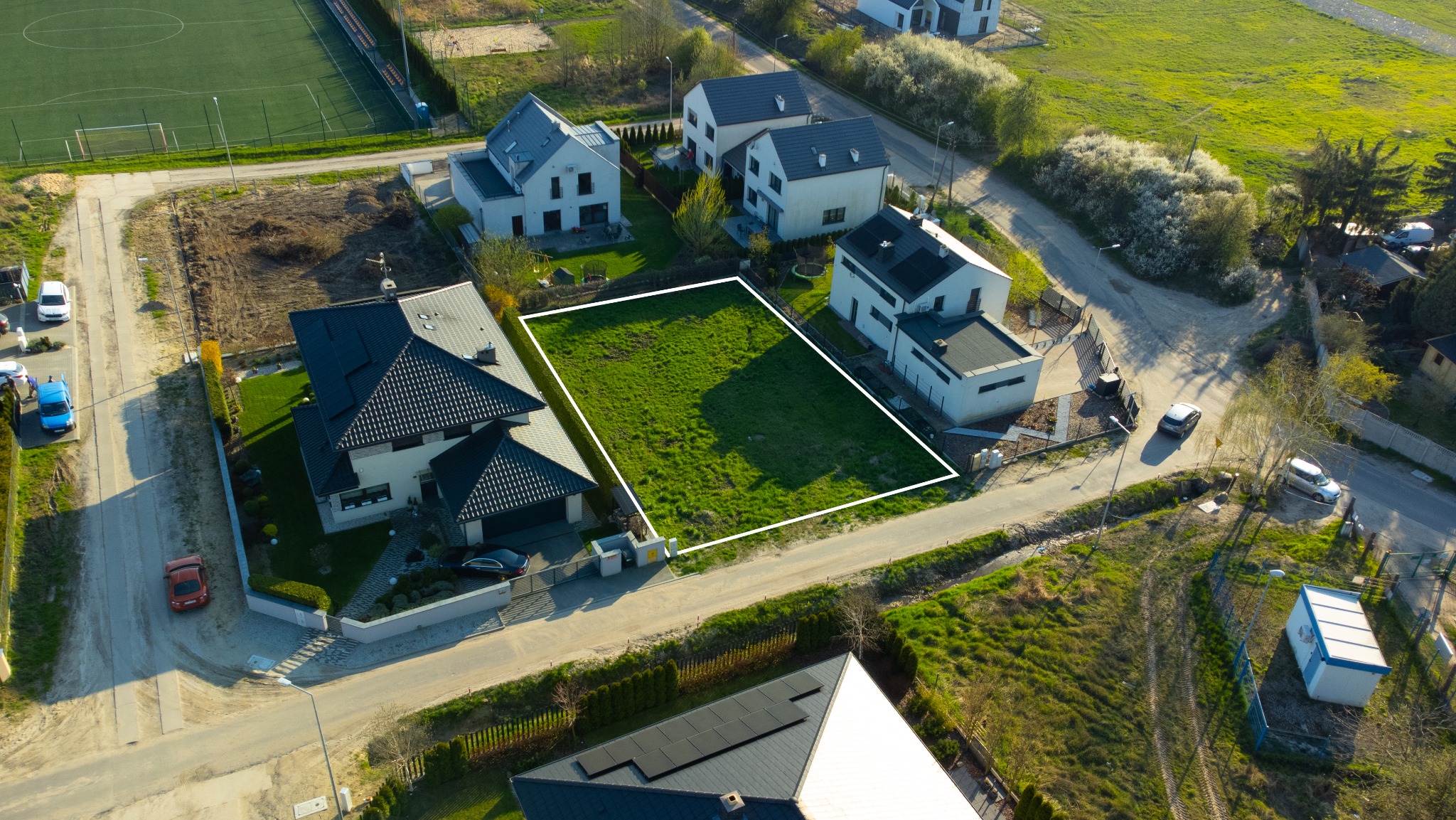 Zdjęcie z lotu ptaka przedstawiające działkę budowlaną zarysowaną białym obrysem, otoczoną nowoczesnymi domami jednorodzinnymi i zielenią, z boiskiem sportowym w tle.