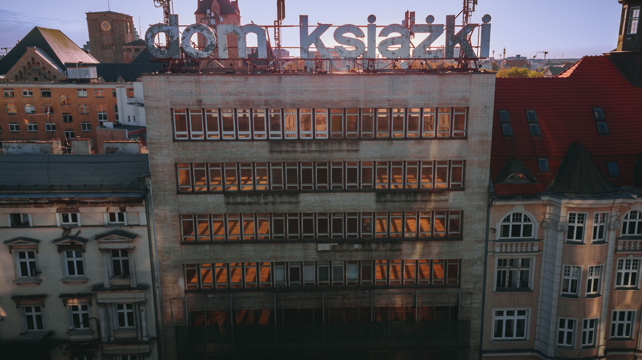 Fasada budynku z napisem 'Dom Książki' na dachu, widok z góry, z odbijającym się w oknach zachodzącym słońcem, otoczona innymi budynkami.