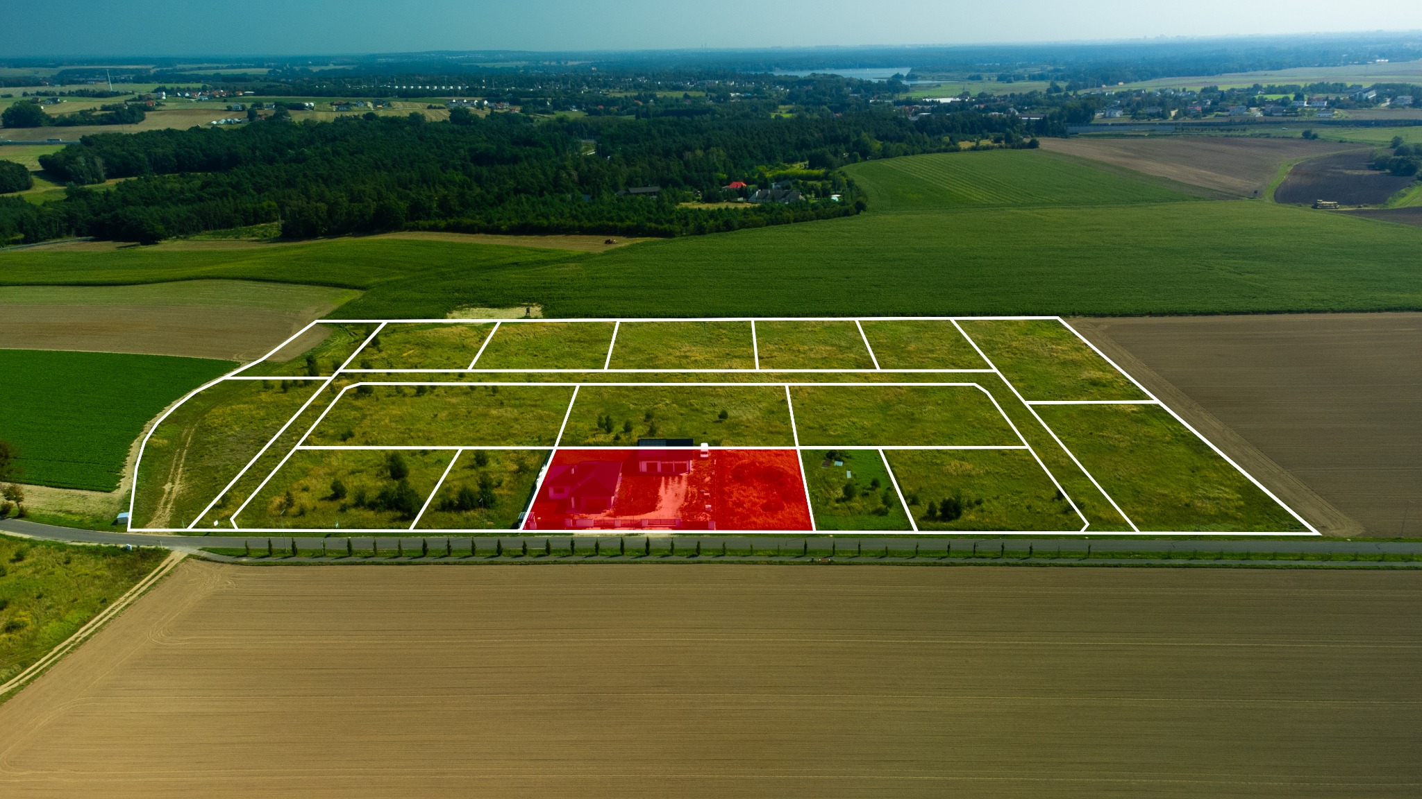 Widok z lotu ptaka na działki budowlane z zaznaczoną na czerwono jedną z nich, otoczone polami i lasami.