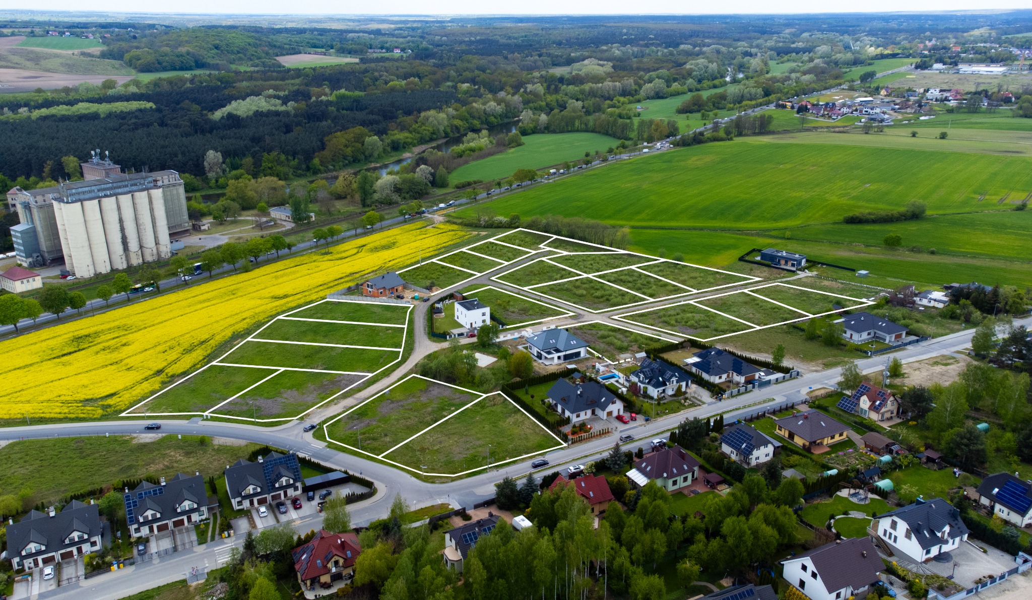 Zdjęcie z drona przedstawia osiedle domów jednorodzinnych z wyznaczonymi działkami na sprzedaż, w tle pola i lasy. Widoczne silosy zbożowe.