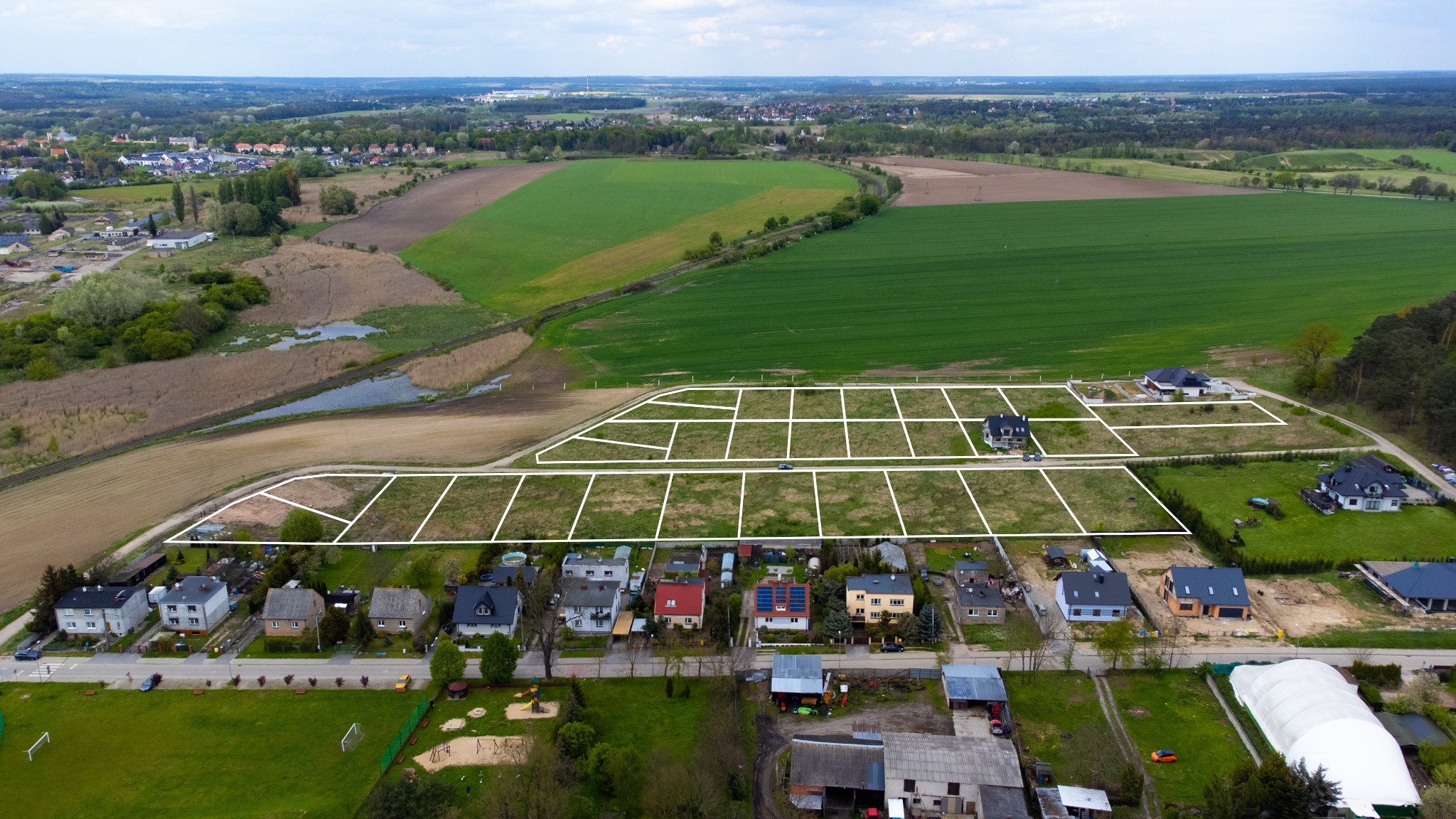 Panoramiczny widok z drona na działki budowlane z naniesionymi granicami, w tle pola uprawne i zabudowa mieszkalna. Ujęcie z góry, słoneczny dzień.