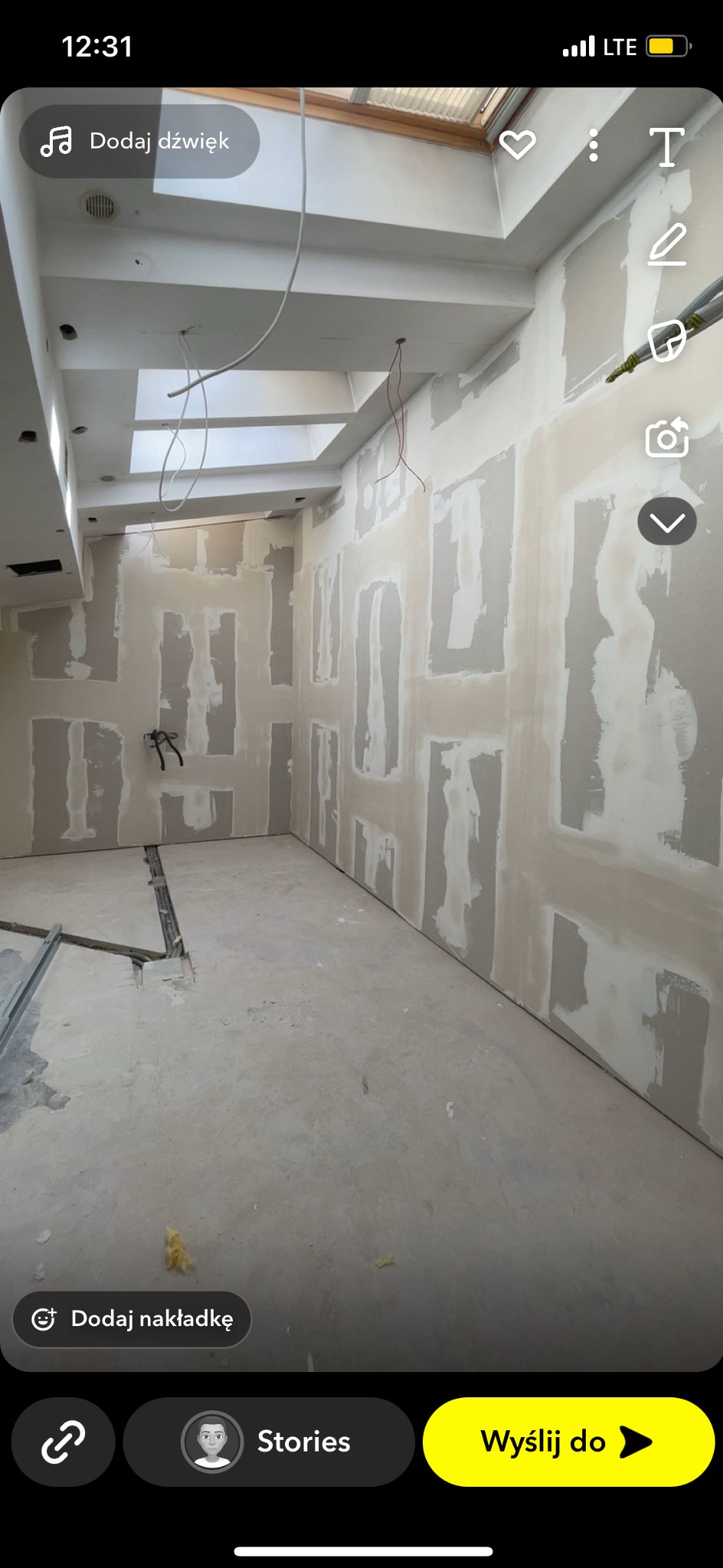 Wnętrze w trakcie prac: ściany z płytami gipsowo-kartonowymi, widoczne łączenia szpachlowane. Na podłodze resztki materiałów budowlanych. Okno dachowe.