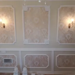 Eleganckie wykończenie ściany z dekoracyjnymi panelami i tapetą w beżowe wzory, oświetlone kinkietami z kryształkami, widoczne elementy sztukaterii i fragment drzwi z czarnymi pasami.