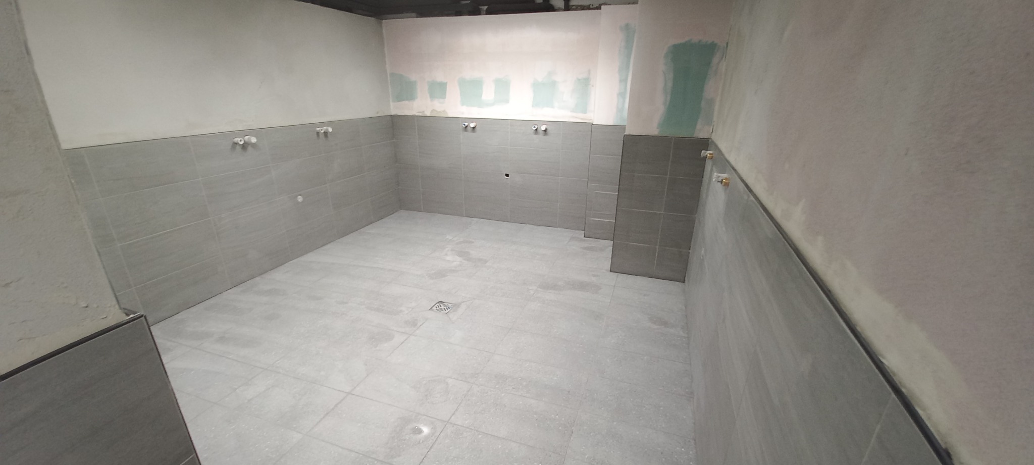 Wnętrze w trakcie prac wykończeniowych: szare płytki na ścianach do połowy wysokości, surowe ściany powyżej z widocznymi śladami szpachlowania, przygotowane punkty instalacji wodnej.