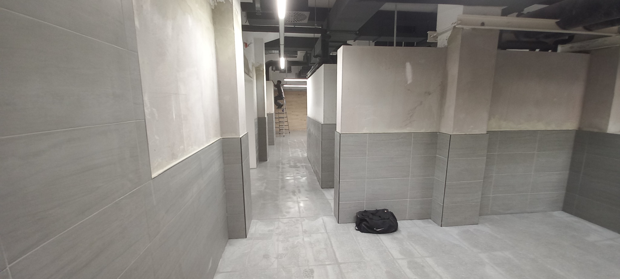 Wnętrze w trakcie prac wykończeniowych: szare płytki ceramiczne na ścianach do połowy wysokości, surowy beton powyżej, widoczny pracownik na drabinie w oddali, czarna torba na podłodze.