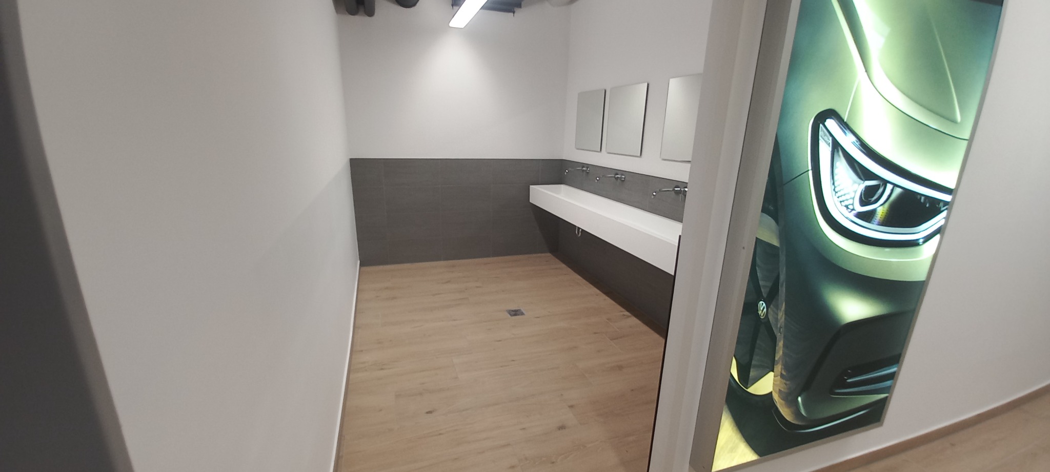 Wnętrze publicznej toalety z trzema lustrami nad długą, białą umywalką, szarą glazurą do połowy ściany i drewnopodobną podłogą, z dodatkowym elementem dekoracyjnym w postaci podświetlonej grafiki...