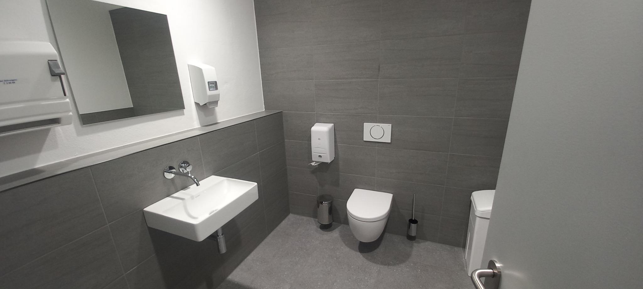 Nowoczesna łazienka w szarościach z wiszącą umywalką, dozownikiem mydła, lustrem i toaletą.