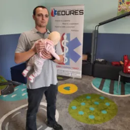 Instruktor z manekinem niemowlęcym prezentuje prawidłowy chwyt podczas kursu pierwszej pomocy dla niemowląt, w tle baner z logo fundacji i kolorowy dywan.