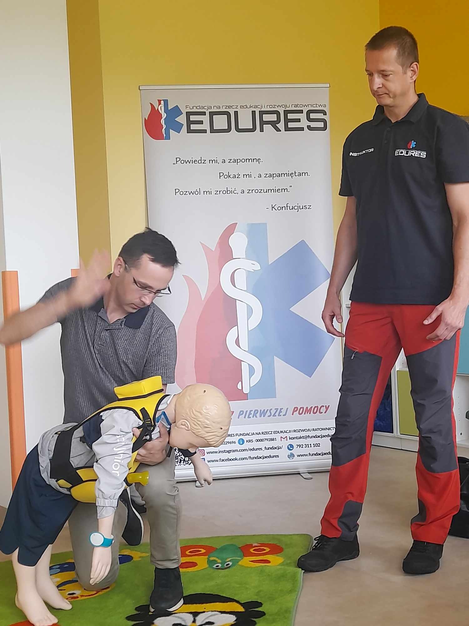 Instruktor demonstruje manewr Heimlicha na fantomie dziecka podczas zajęć, drugi instruktor obserwuje, w tle baner z logo i hasłami edukacyjnymi.