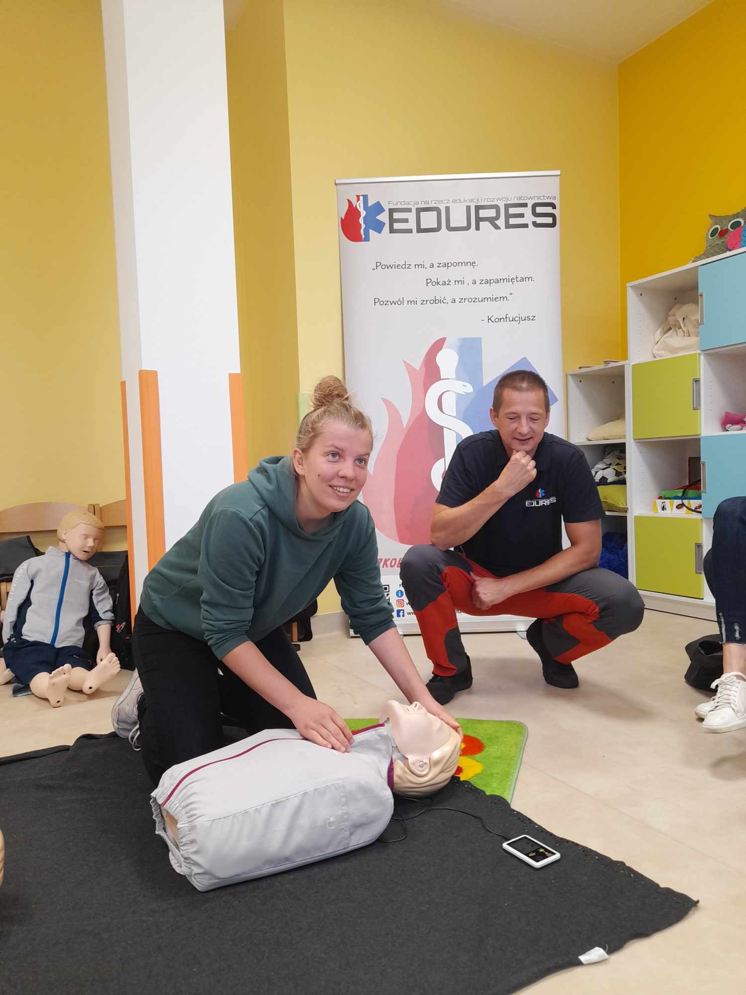 Kobieta ćwiczy resuscytację krążeniowo-oddechową na fantomie, obok instruktor demonstruje prawidłową pozycję, w tle baner edukacyjny oraz manekin.