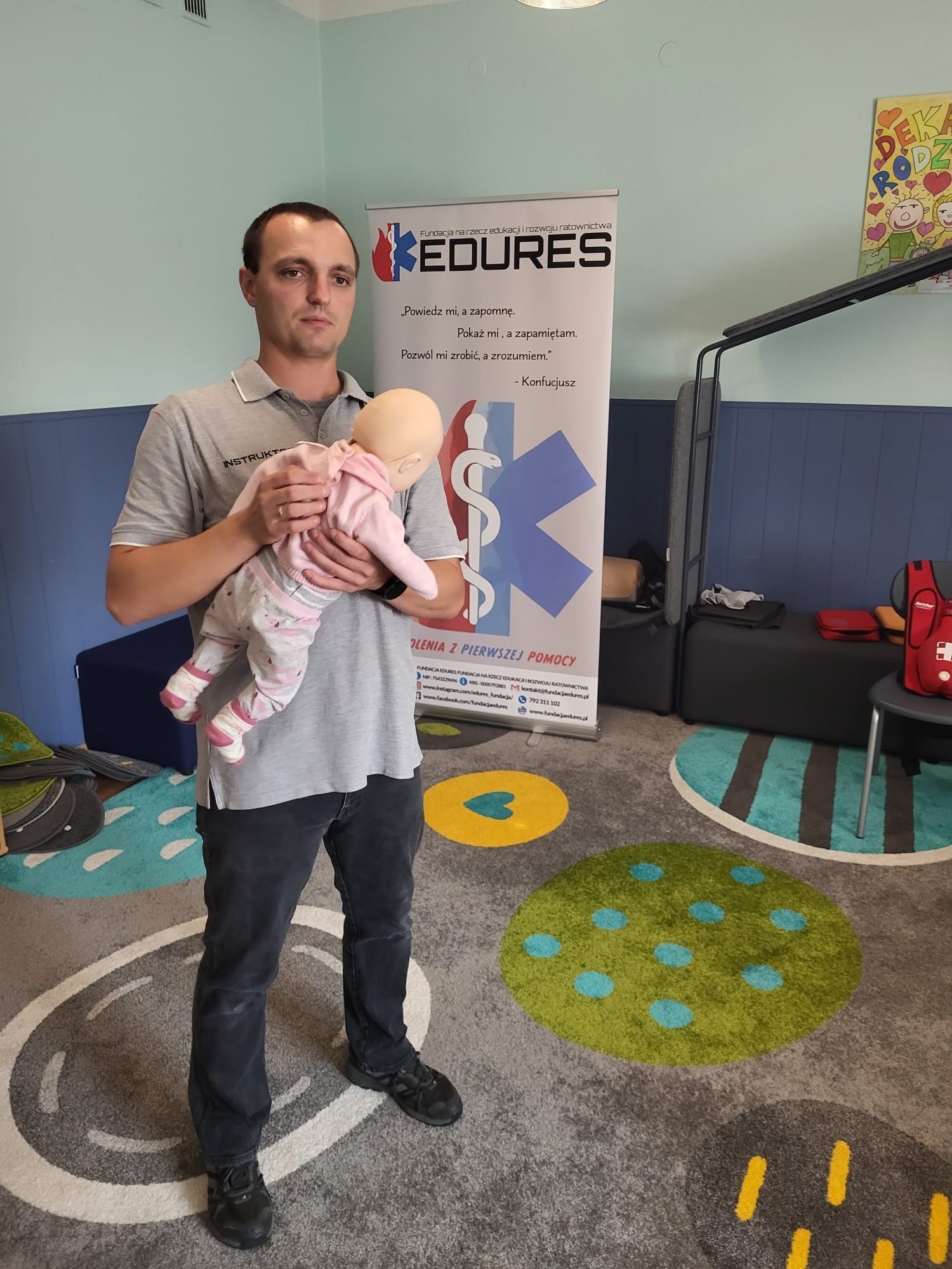 Instruktor z manekinem niemowlęcym prezentuje prawidłowy chwyt podczas kursu pierwszej pomocy dla niemowląt, w tle baner z logo fundacji i kolorowy dywan.