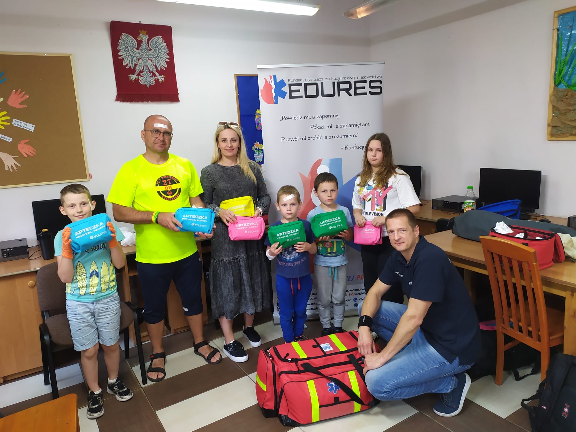 Grupa osób, w tym dzieci z opatrunkami, prezentuje kolorowe apteczki pierwszej pomocy przed banerem edukacyjnym EDURES.