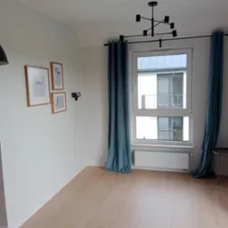 Jasne wnętrze pokoju z drewnianą podłogą, białymi ścianami, trzema obrazkami w drewnianych ramkach, nowoczesną lampą sufitową i turkusowymi zasłonami.