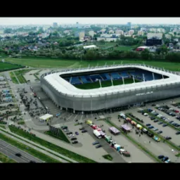 Panoramiczny widok z lotu ptaka na Arena Lublin, otoczony parkingami i zielenią, z widoczną grupą ludzi i pojazdów, w tle zabudowa miejska.