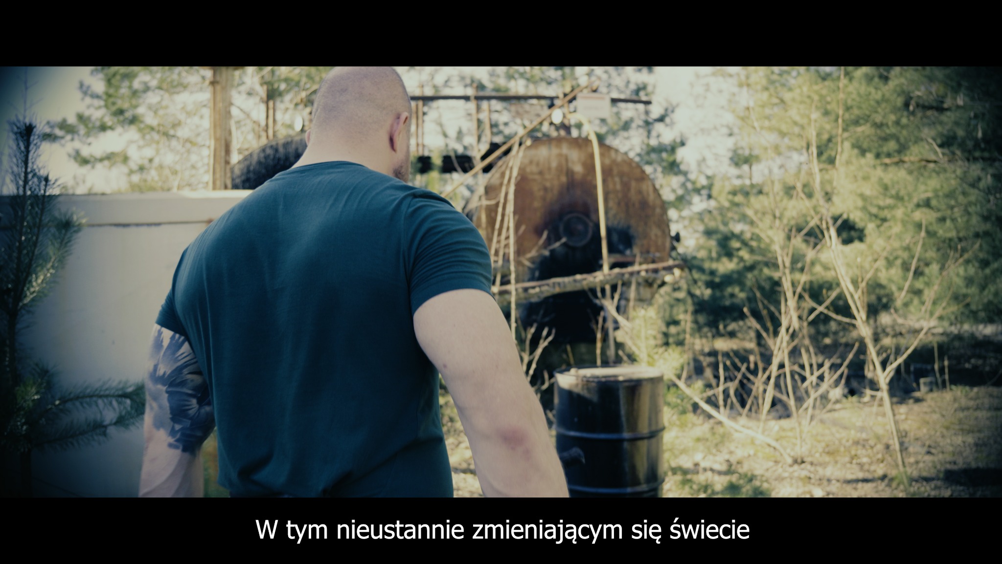 Mężczyzna w zielonej koszulce z tatuażem na ramieniu, stojący tyłem do kamery, na tle zardzewiałej konstrukcji i lasu, fragment ujęcia z filmu.
