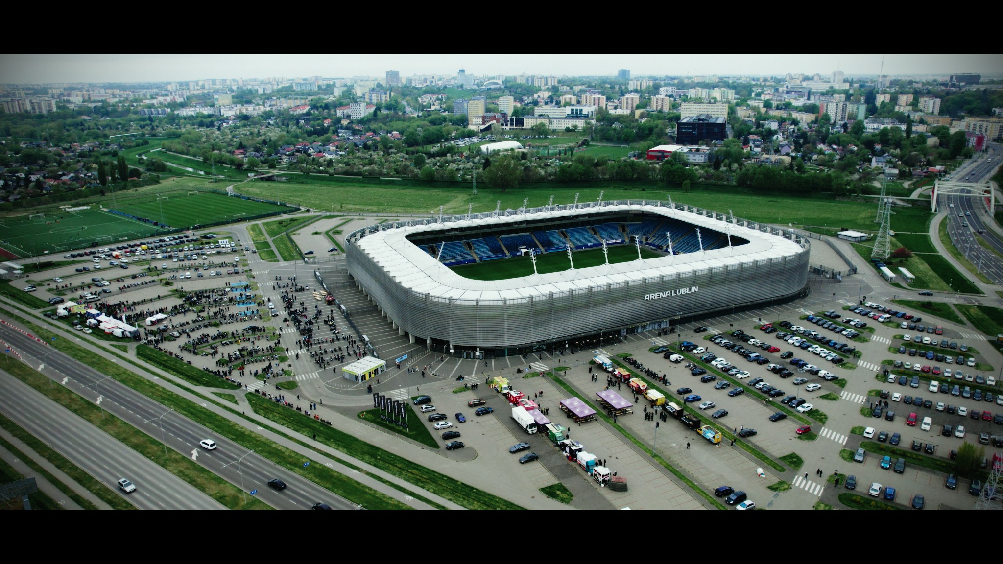 Panoramiczny widok z lotu ptaka na Arena Lublin, otoczony parkingami i zielenią, z widoczną grupą ludzi i pojazdów, w tle zabudowa miejska.