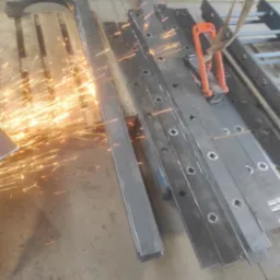 Cięcie metalowych elementów konstrukcyjnych z iskrami, widoczne profile z kwadratowymi otworami i gięty element na palecie, uchwyt transportowy z pasem.