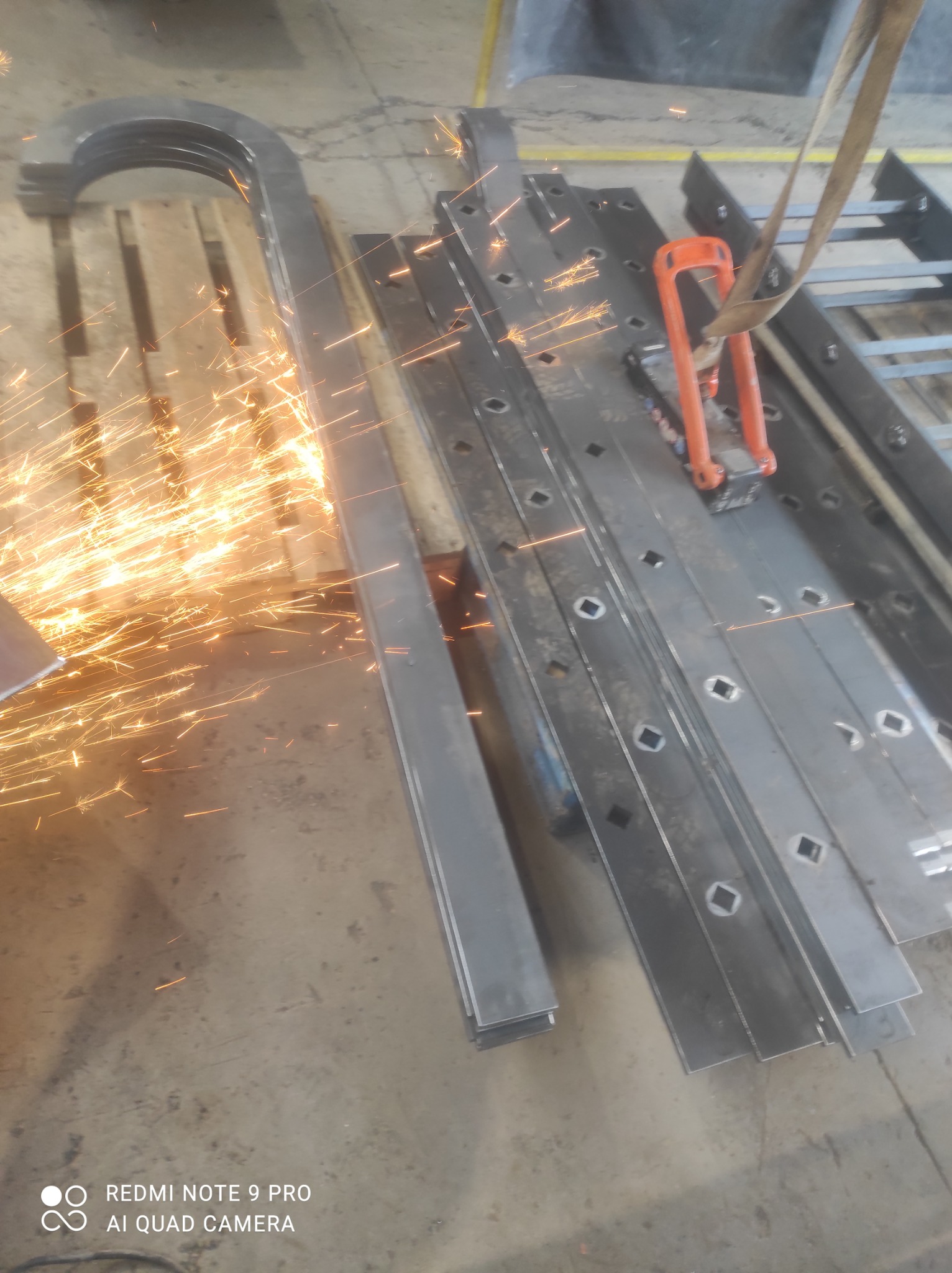 Cięcie metalowych elementów konstrukcyjnych z iskrami, widoczne profile z kwadratowymi otworami i gięty element na palecie, uchwyt transportowy z pasem.