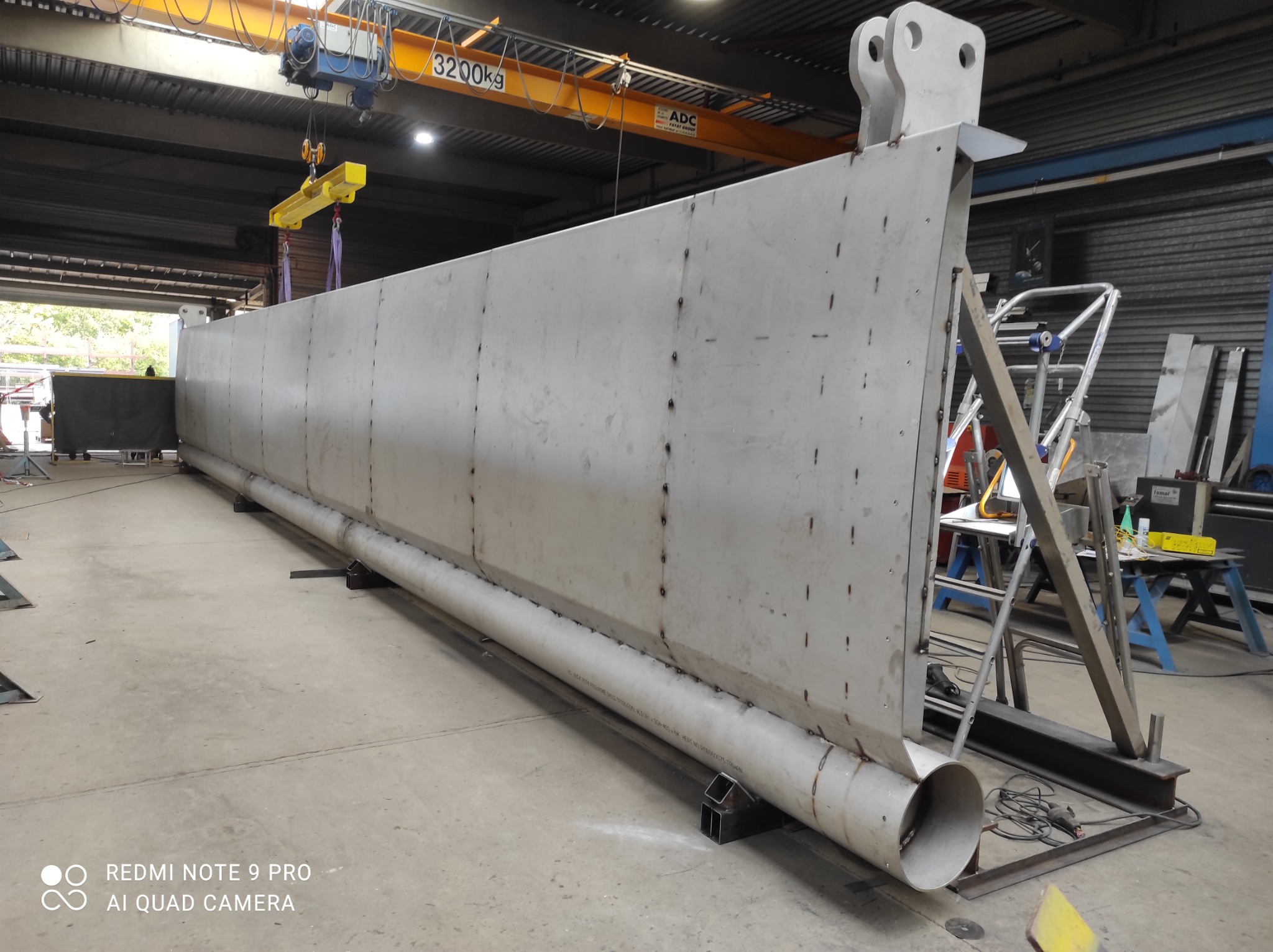 Długa, spawana konstrukcja stalowa w hali produkcyjnej, podnoszona suwnicą z napisem 3200kg, widoczne spawane elementy i rura wzdłuż dolnej krawędzi.