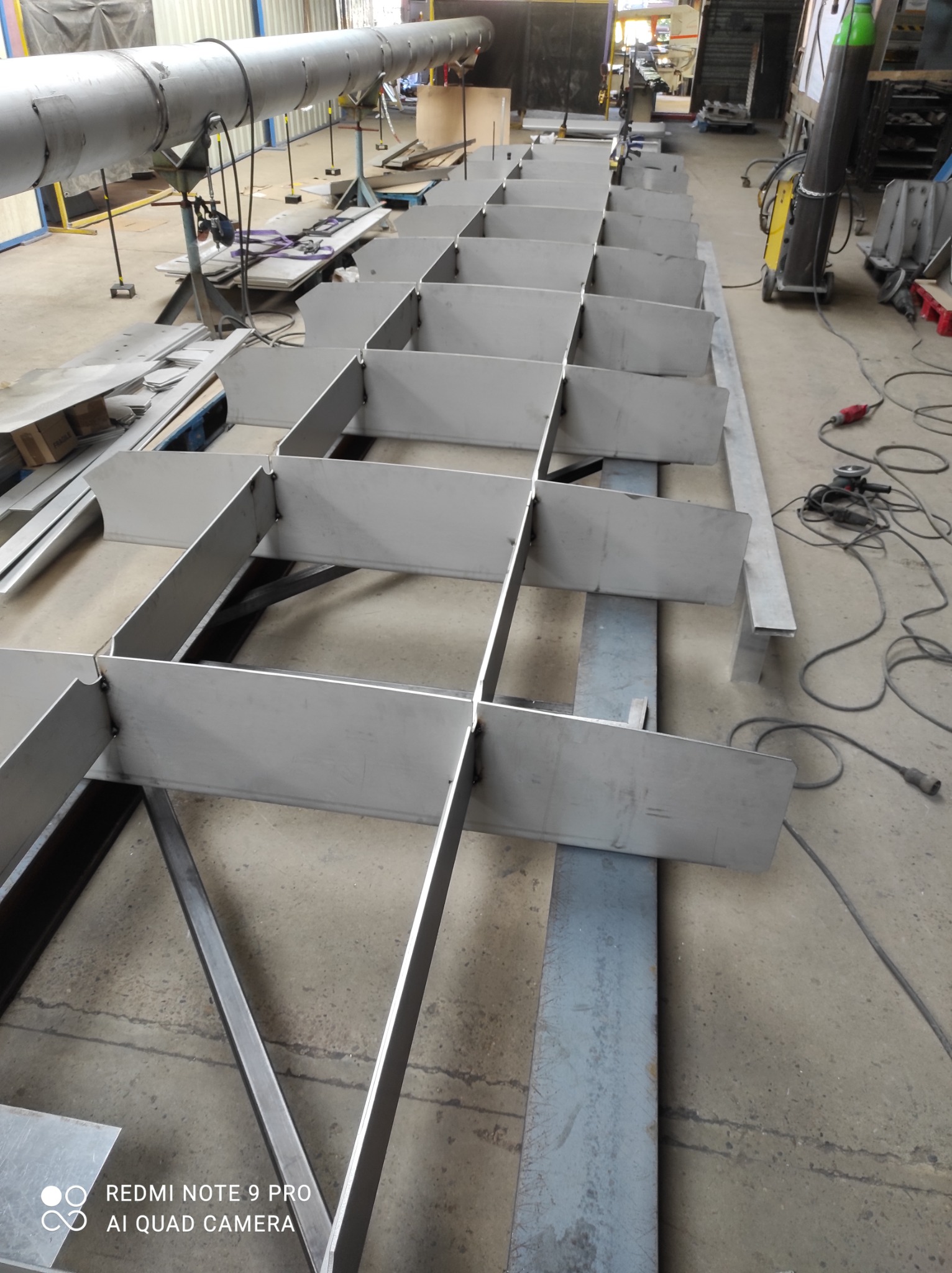 Wnętrze hali produkcyjnej z widoczną konstrukcją stalową w trakcie budowy, spawana kratownica z profili stalowych i blach, narzędzia spawalnicze w tle.