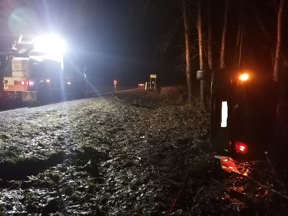 Nocne zdjęcie przewróconego samochodu osobowego na poboczu drogi, oświetlonego światłami awaryjnymi i reflektorami pojazdu pomocy drogowej w oddali.