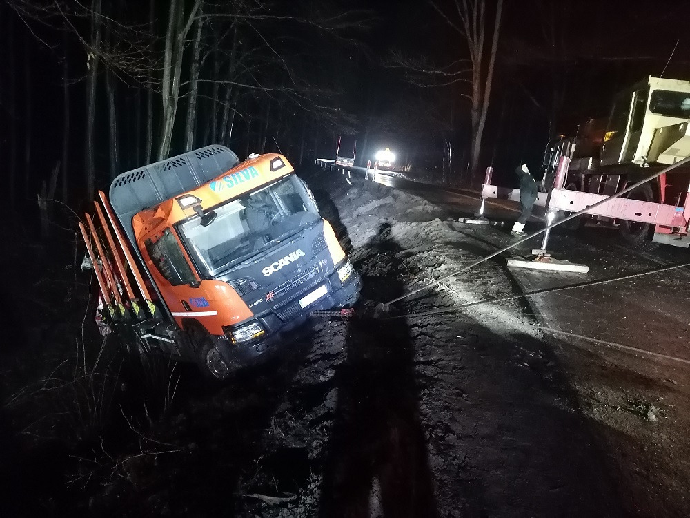Pomarańczowa ciężarówka Scania częściowo poza drogą, przechylona w błocie, holowana linami przez dużą, żółtą ciężarówkę z wysięgnikiem w ciemnościach, widoczny cień osoby.
