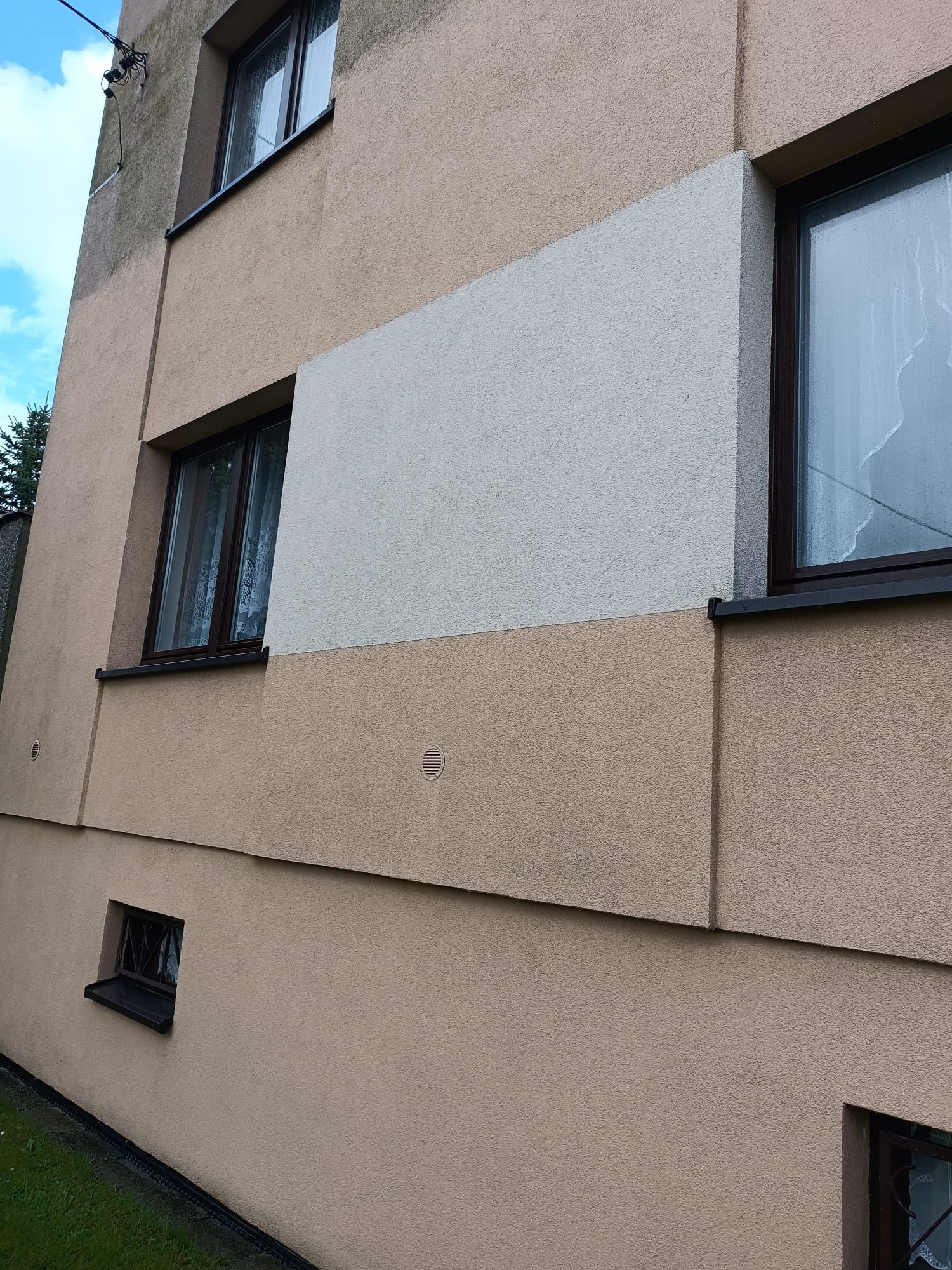 Fragment elewacji budynku wielorodzinnego z oknami, po częściowym odświeżeniu koloru, gdzie widać różnicę między starą a nową, jaśniejszą farbą.