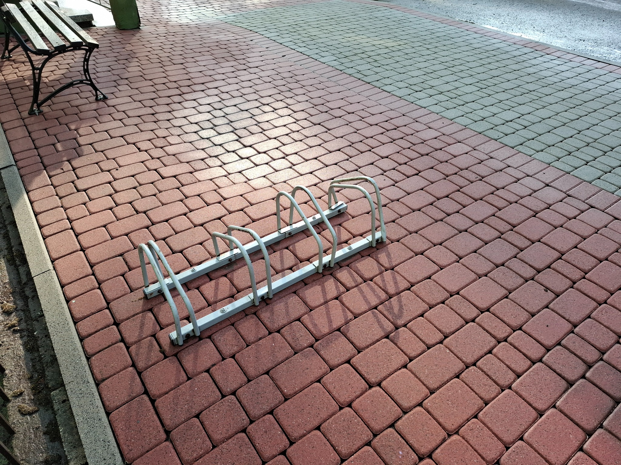 Czerwono-szara kostka brukowa z metalowym stojakiem na rowery i ławką w tle.