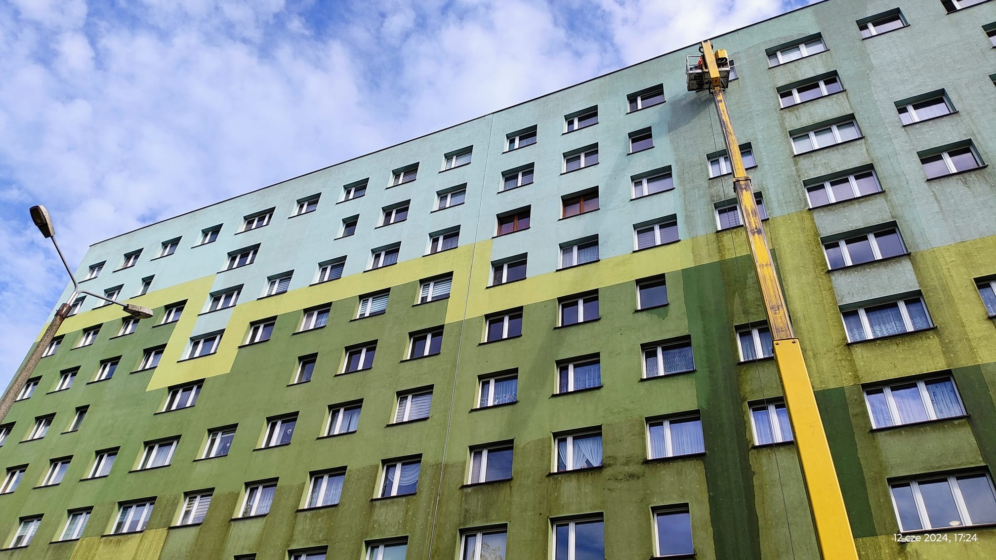 Wielopiętrowy budynek pomalowany w odcieniach zieleni i błękitu, czyszczony z wykorzystaniem podnośnika koszowego, widoczne ślady po czyszczeniu na elewacji.