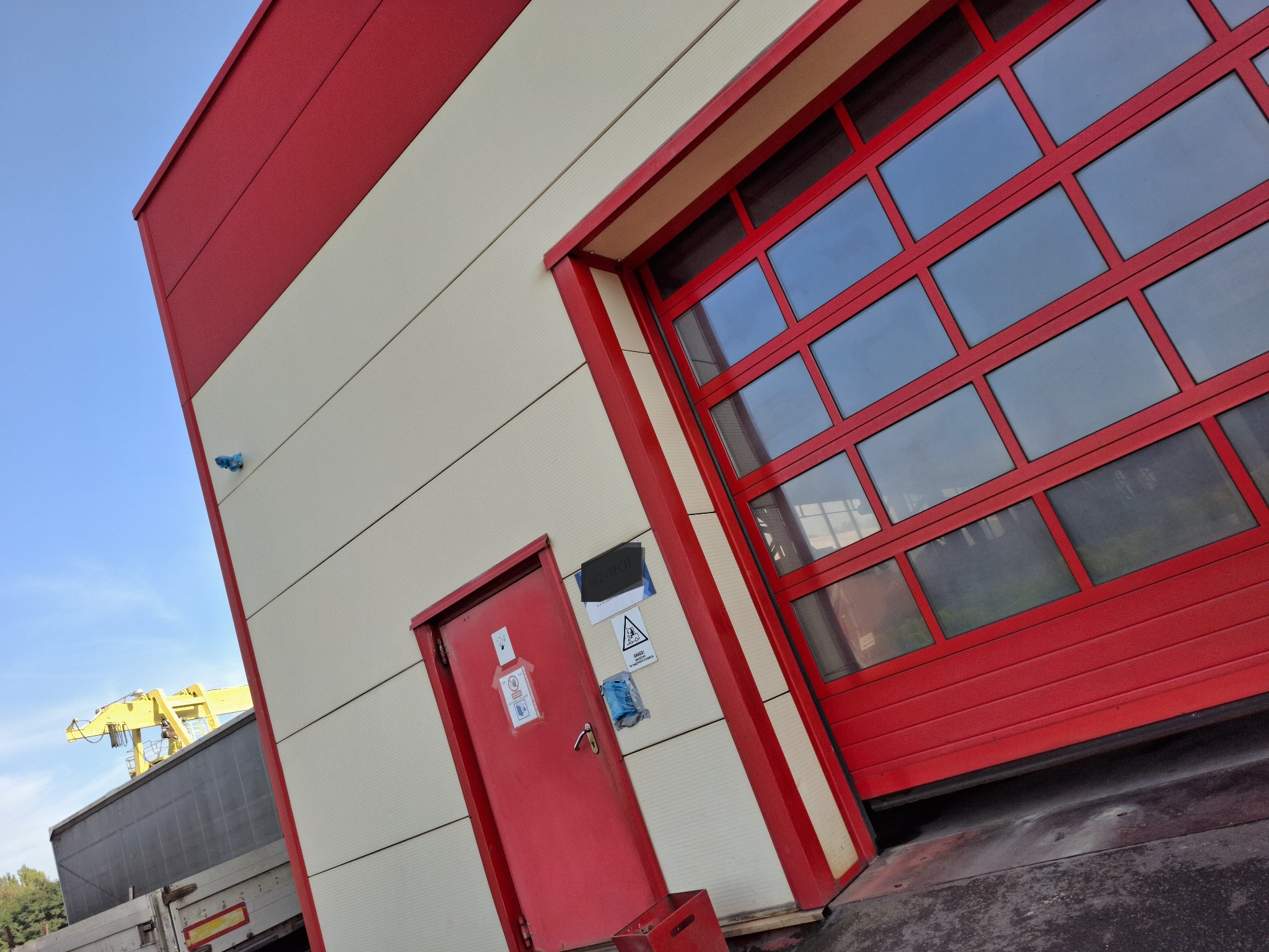 Czerwono-biała elewacja budynku przemysłowego z czerwoną bramą segmentową i drzwiami wejściowymi, widoczny fragment żurawia w tle.