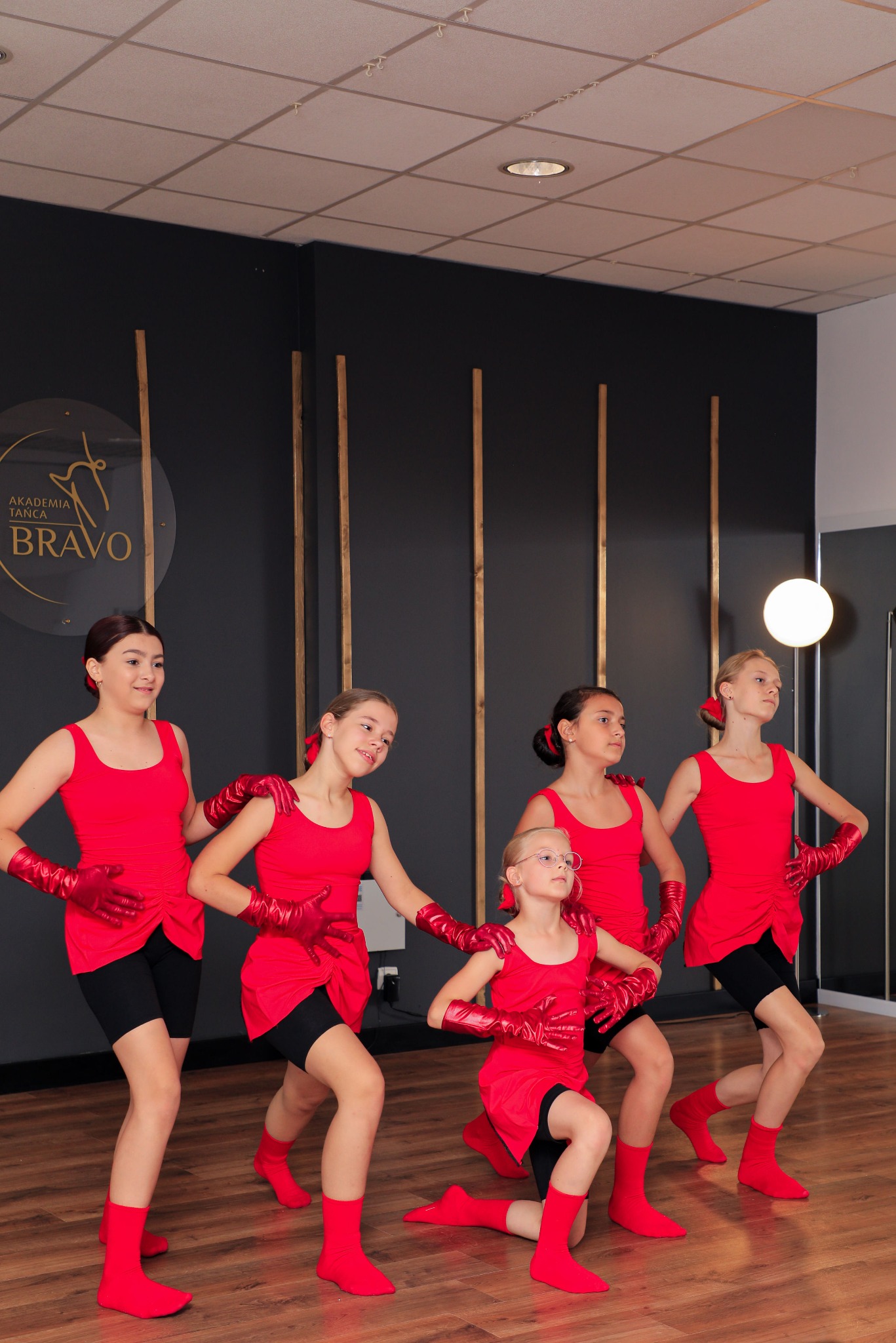 Grupa dziewcząt w czerwonych kostiumach i rękawiczkach prezentuje choreografię taneczną w sali z drewnianą podłogą i logo Akademii Tańca Bravo na ścianie.