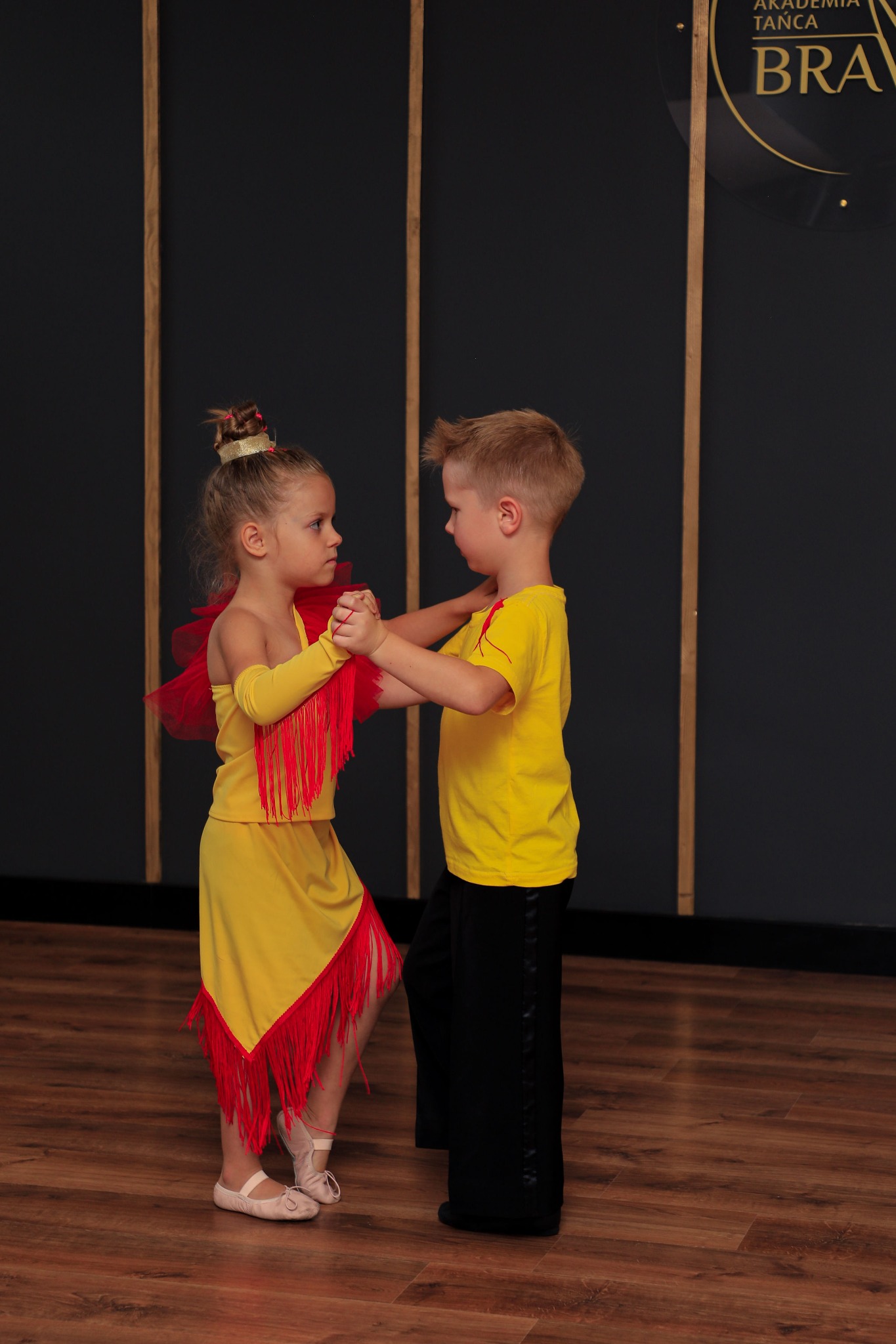 Dwoje dzieci w strojach tanecznych, dziewczynka w żółtej sukience z czerwonymi frędzlami, chłopiec w żółtej koszulce i czarnych spodniach, trzymają się za ręce na tle ciemnej ściany z logo szkoły...
