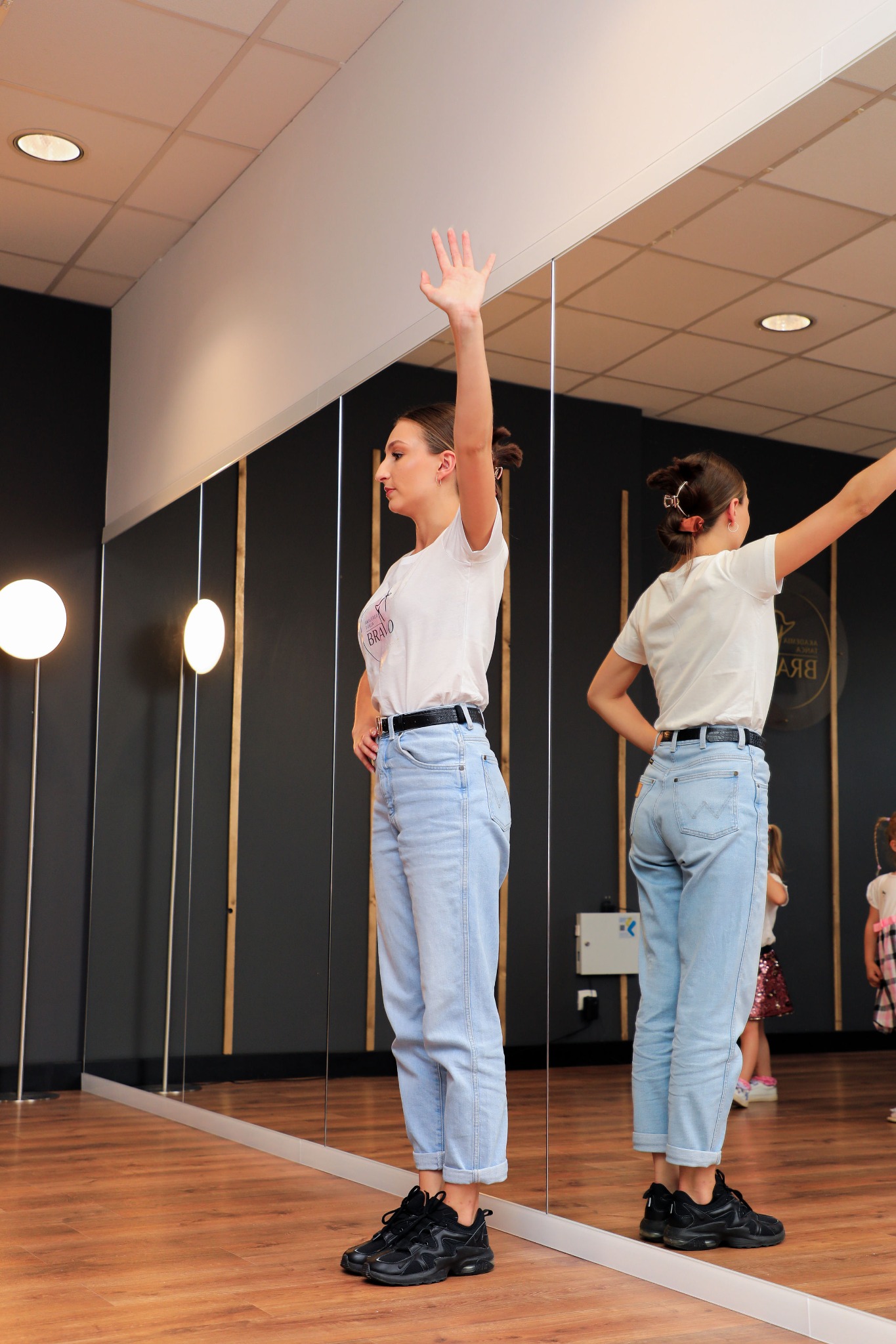 Instruktorka w jasnych jeansach i białej koszulce z podniesioną ręką w sali tanecznej z lustrami, widoczne dzieci w tle.