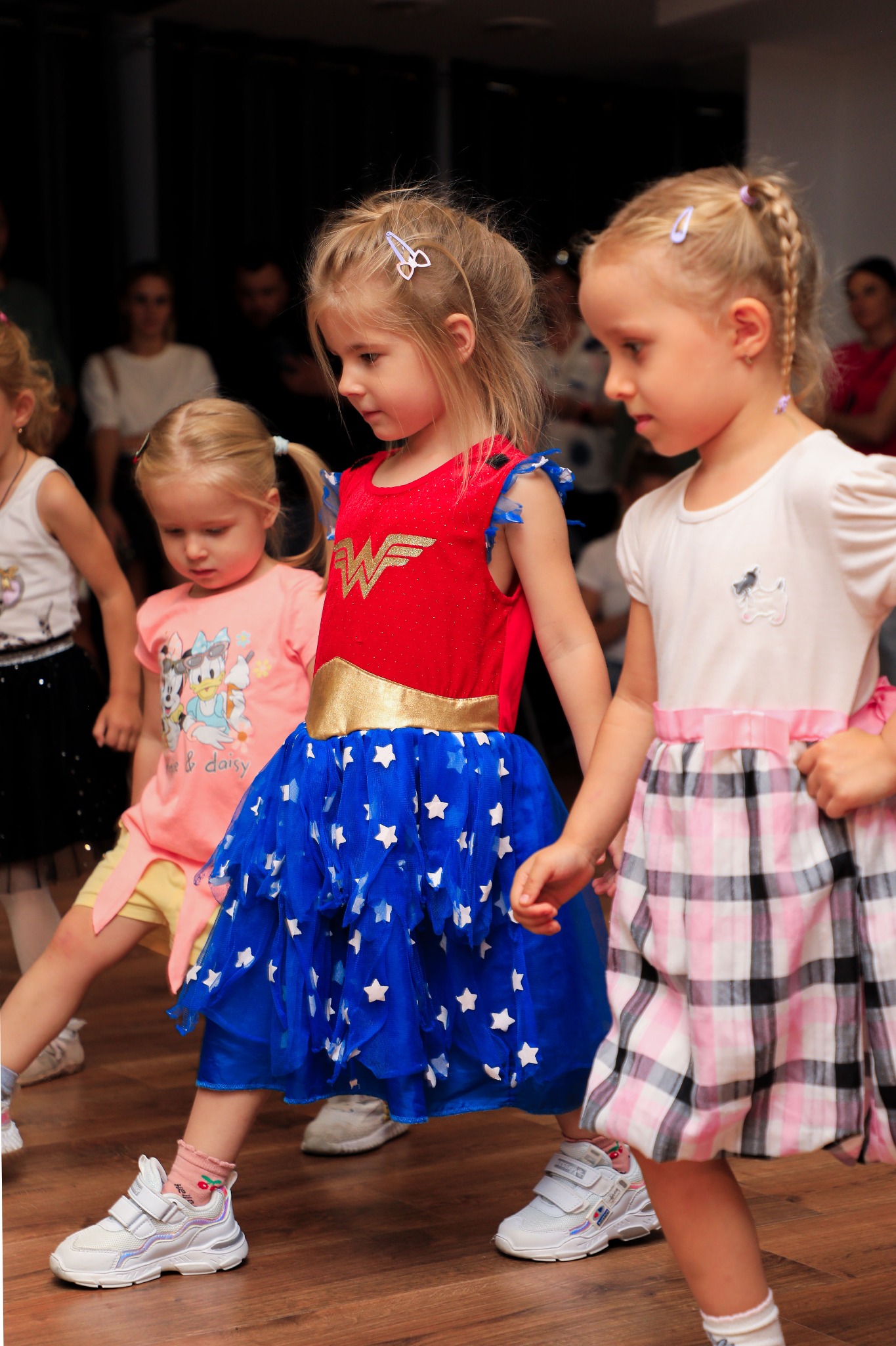 Grupa dziewczynek w kostiumach, w tym jedna w stroju Wonder Woman, podczas zajęć tanecznych na drewnianym parkiecie.