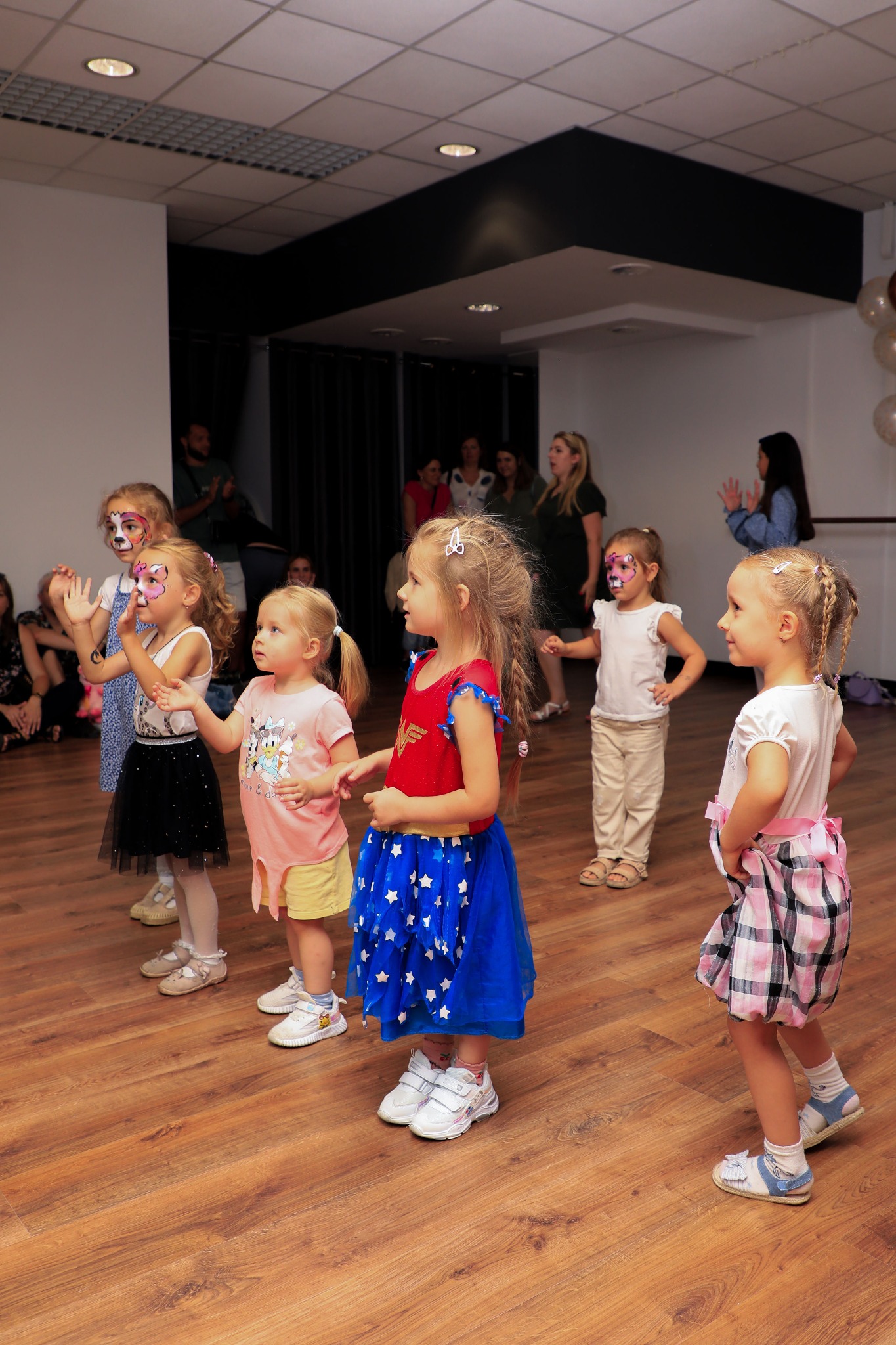 Grupa dziewczynek z pomalowanymi twarzami w kostiumach, ustawionych w rzędzie na drewnianej podłodze, ćwiczących układ taneczny w jasnym pomieszczeniu z rodzicami w tle.
