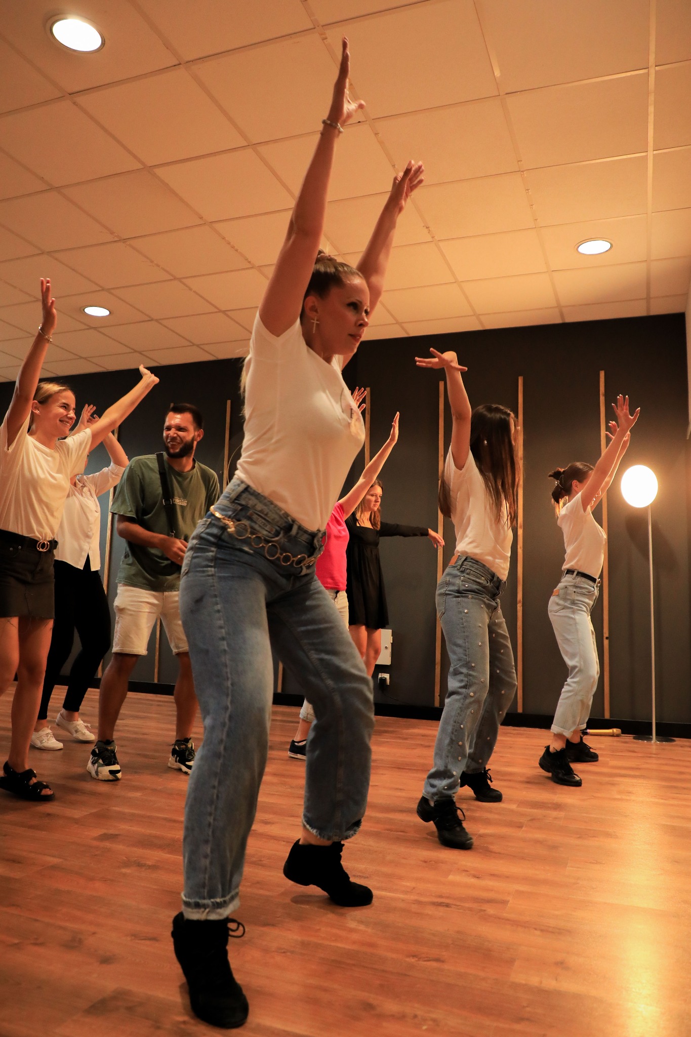 Grupa osób w jasnych ubraniach i jeansach ćwiczy układ taneczny w przestronnym, dobrze oświetlonym studio z drewnianą podłogą i ciemną ścianą w tle.