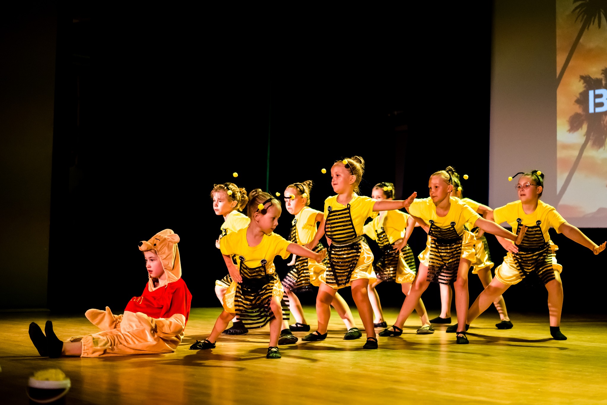 Grupa dziewcząt w kostiumach pszczół i misia Puchatka prezentuje układ taneczny na scenie z ciemnym tłem i projekcją palm.