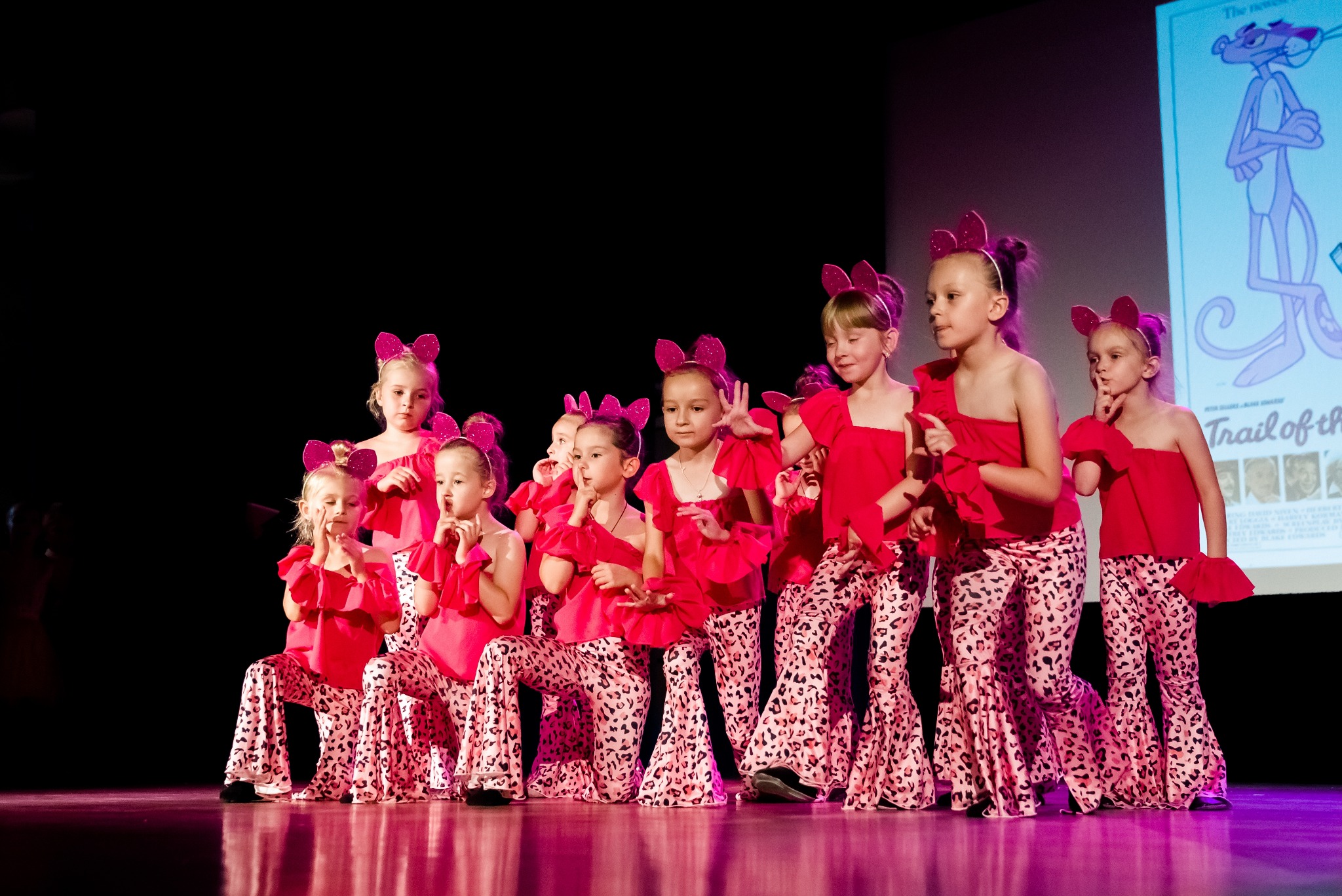 Grupa dziewczynek w czerwonych strojach i uszach z kokardami, pozuje na scenie z projekcją Różowej Pantery w tle, podczas występu tanecznego.
