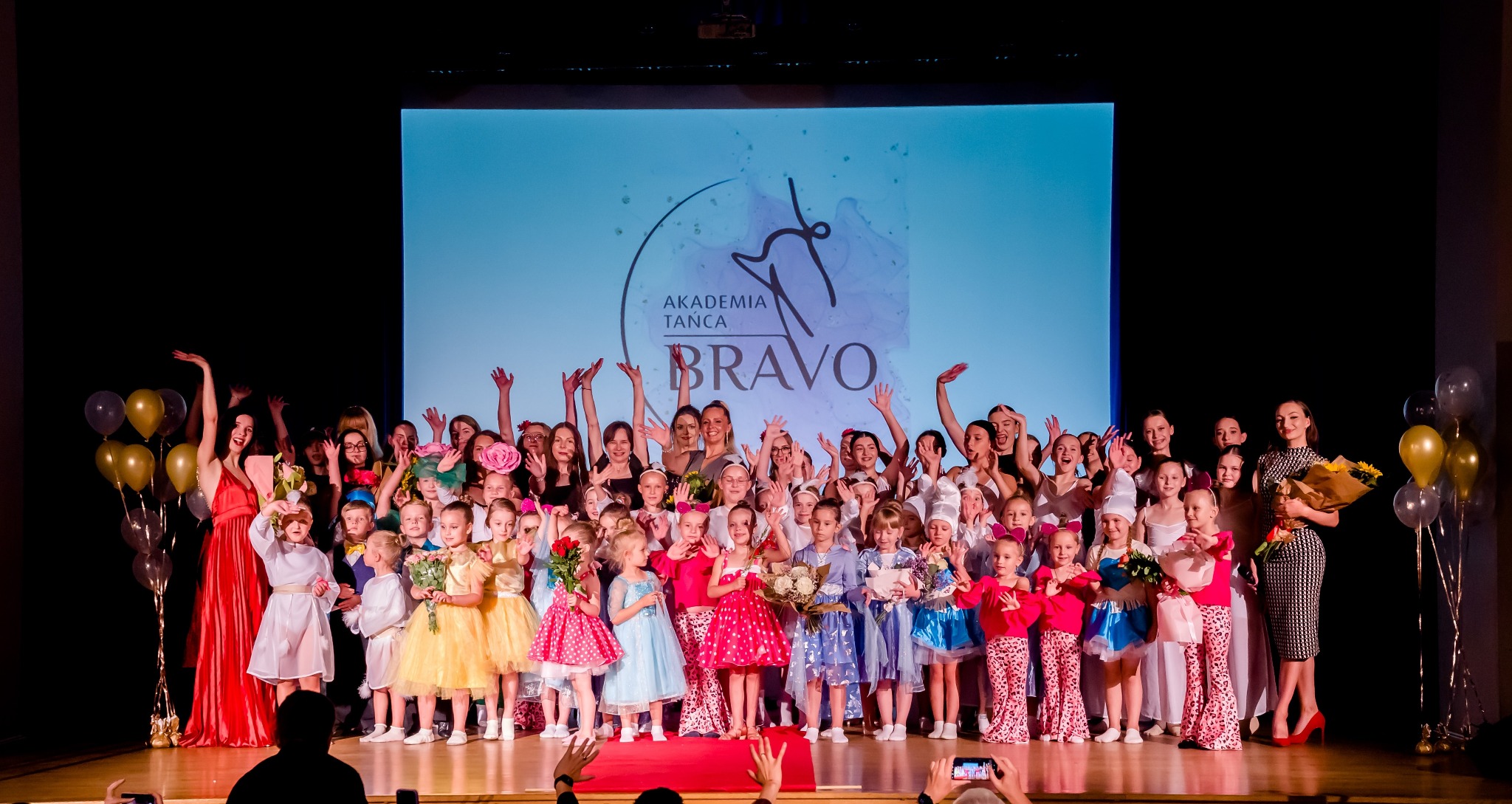 Grupowe zdjęcie uczniów i instruktorów Akademii Tańca Bravo na scenie po występie, z logo szkoły wyświetlonym w tle, a uczestnicy trzymają bukiety kwiatów i balony.