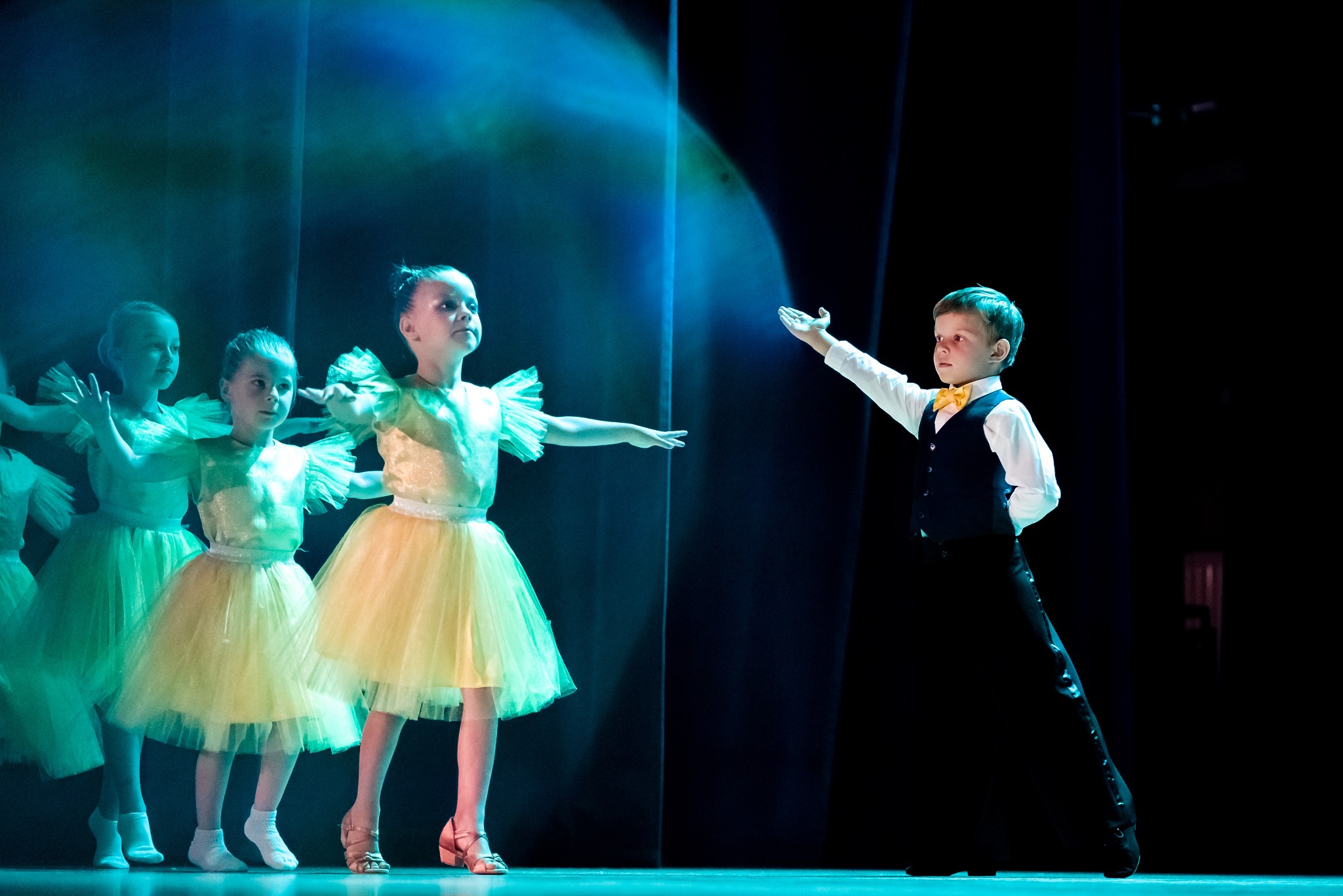 Grupa dzieci w kostiumach tanecznych na scenie podczas występu, oświetlona reflektorami; dziewczynki w żółto-zielonych sukienkach i chłopiec w czarnej kamizelce i muszce.