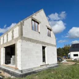 Surowy stan domu z białych bloczków, widoczne otwory okienne bez ram, zarys fundamentów, w tle drugi dom z panelami słonecznymi na dachu, błękitne niebo z obłokami.