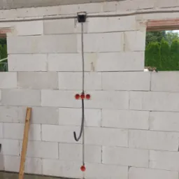 Surowa ściana z betonu komórkowego w trakcie budowy, z widoczną instalacją elektryczną: puszką przy suficie, kablem zwisającym w dół i trzema czerwonymi puszkami podtynkowymi na wysokości gniazdek.