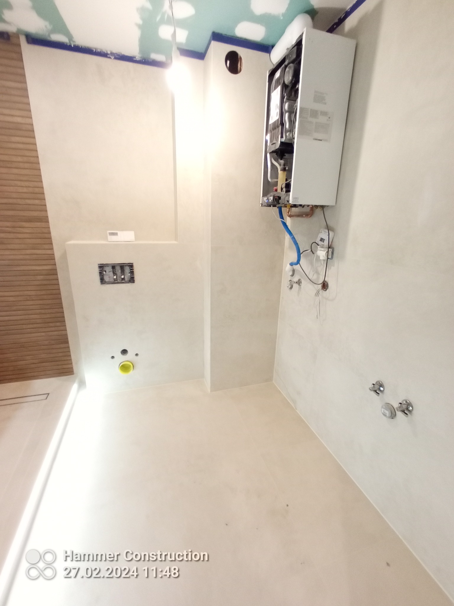 Nowa łazienka w trakcie prac wykończeniowych: widoczny zamontowany piec gazowy, przygotowane przyłącza wodne, gładkie ściany w jasnym odcieniu, częściowo wykończony sufit i fragment drewnianej...