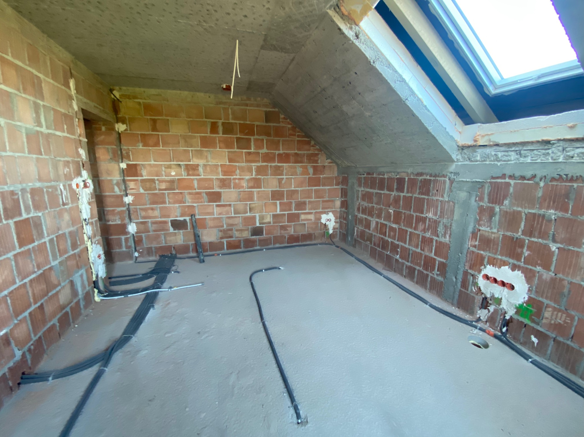 Surowy stan poddasza z widocznymi przewodami elektrycznymi ułożonymi na podłodze i wyprowadzonymi ze ściany, ceglana ściana, okno dachowe.