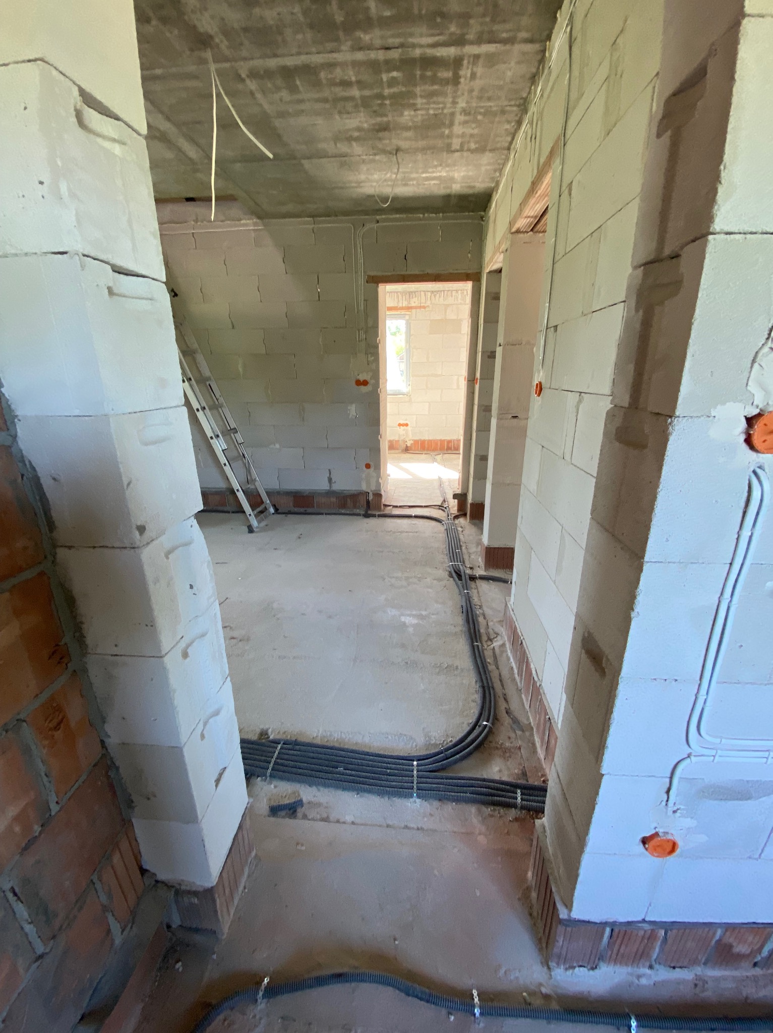 Wnętrze surowego domu w Głogowie Małopolskim w trakcie prac instalacyjnych, widoczne kable w peszlach ułożone wzdłuż ścian, puszki elektryczne w ścianach z betonu komórkowego, drabina oparta o ścianę.