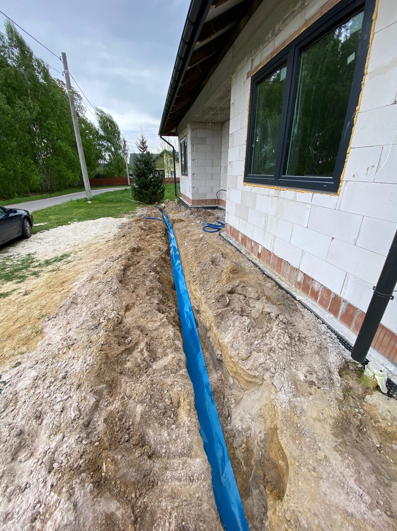 Wykonany wykop pod przyłącze elektryczne do budynku z białego bloczka, zabezpieczony niebieską folią ochronną, widok z góry na teren budowy w pochmurny dzień.