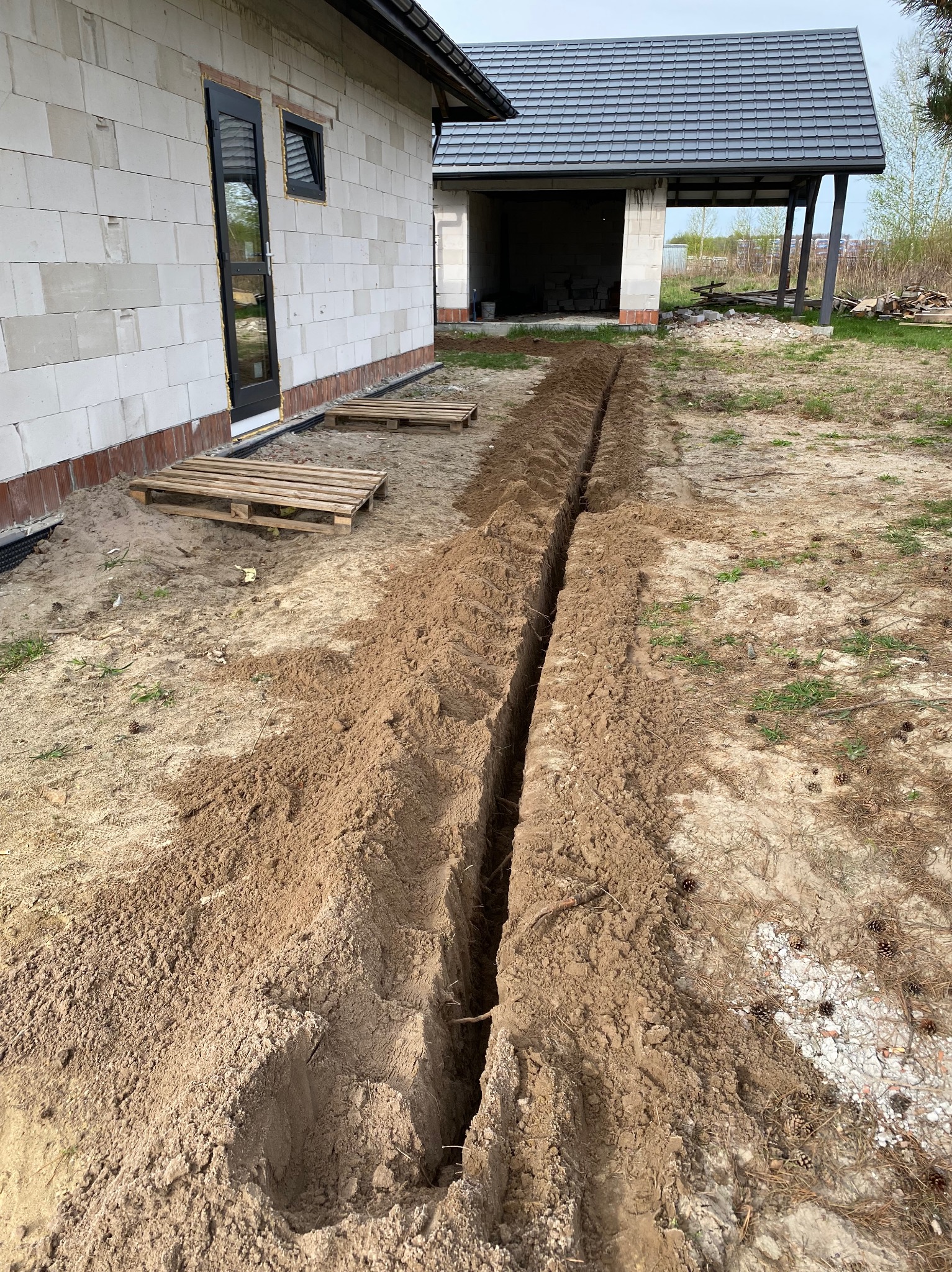 Wykopany wąski rów w ziemi między dwoma budynkami w trakcie budowy, prawdopodobnie pod instalację kablową.