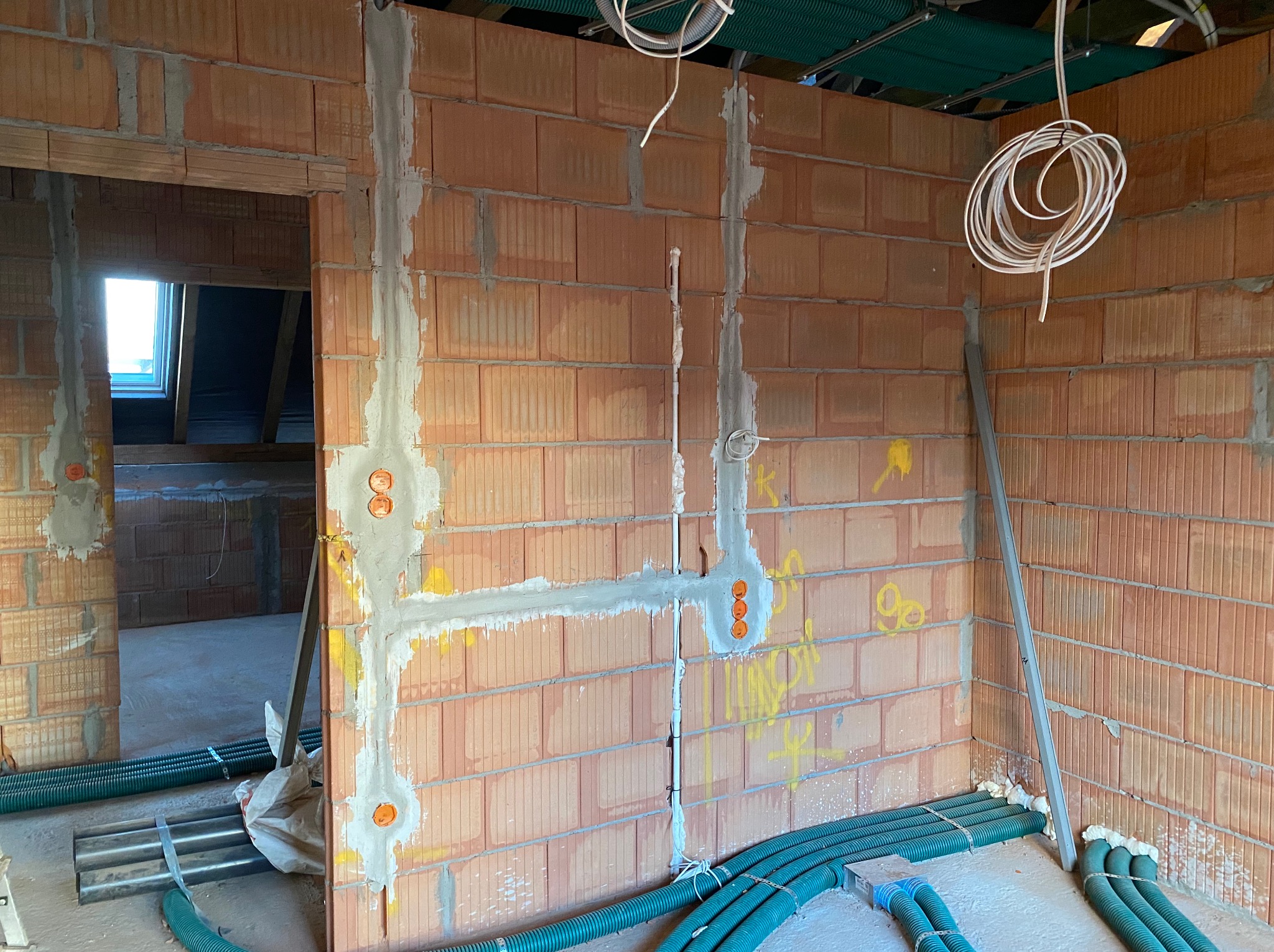 Surowa ściana z czerwonej cegły z wyżłobieniami wypełnionymi zaprawą pod przewody elektryczne i puszkami, zielone rury karbowane na podłodze, widok na poddasze przez otwór drzwiowy.