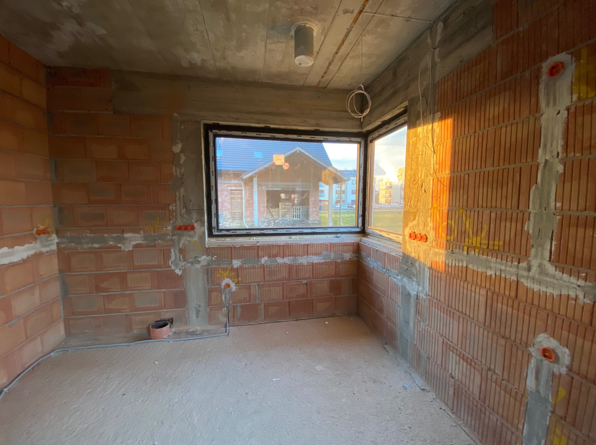 Surowy stan wnętrza z cegły ceramicznej, widoczne puszki instalacyjne w ścianach, okno z widokiem na budynek w budowie, betonowy strop z wystającymi przewodami.